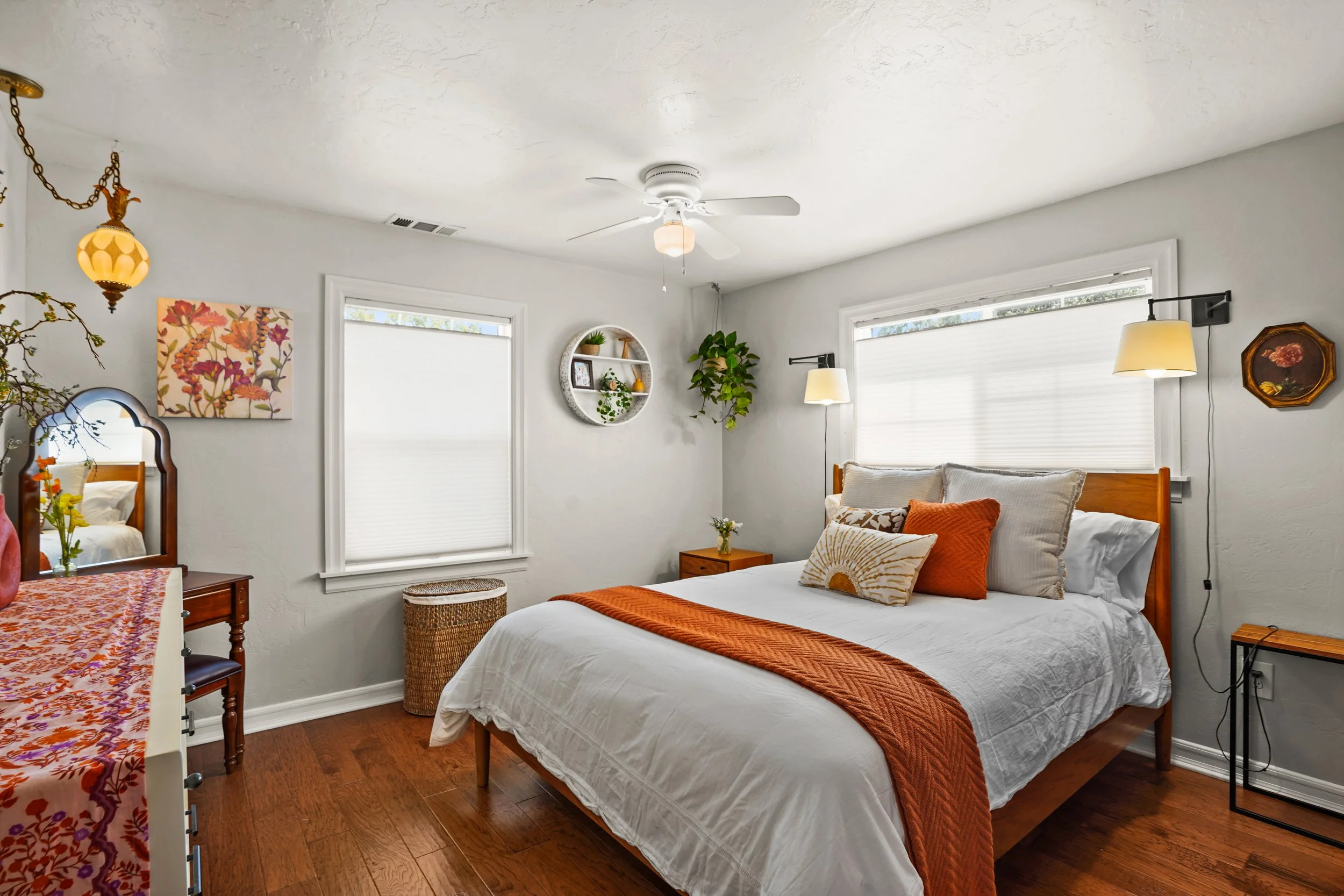 A cozy bedroom with white walls and wooden flooring. There is a white bed with a rust-colored blanket and matching pillows. The room has large windows with white blinds, a ceiling fan with a light, and various wall decorations including a floral pain
