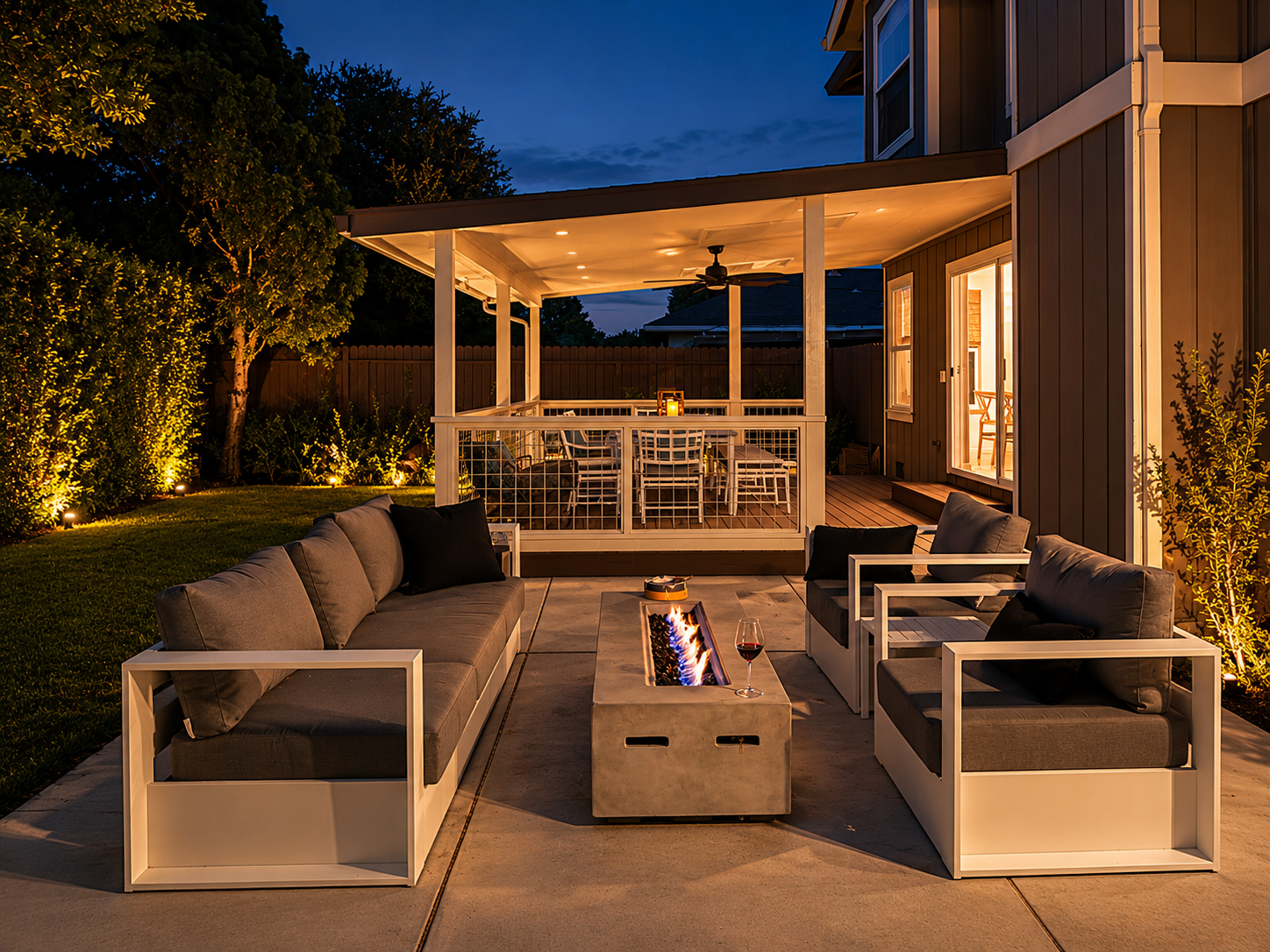 Backyard patio at night with seating area, fire pit, and outdoor dining space illuminated by warm lighting.