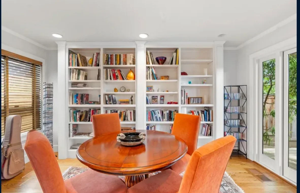 Dining room with round wooden table, four orange upholstered chairs, built-in white bookshelves filled with books and decorative items, hardwood floor, window with wooden blinds, and glass doors with greenery outside.