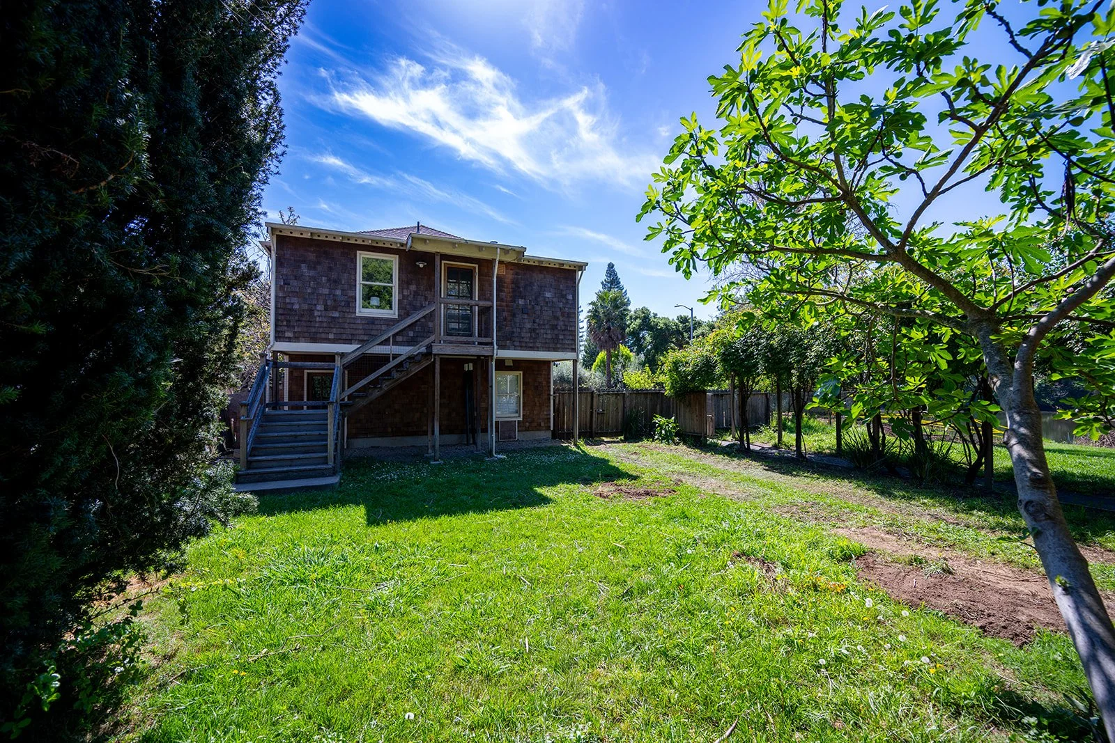 Backyard view of a house with a wooden staircase, surrounded by green grass, trees, and a clear blue sky.