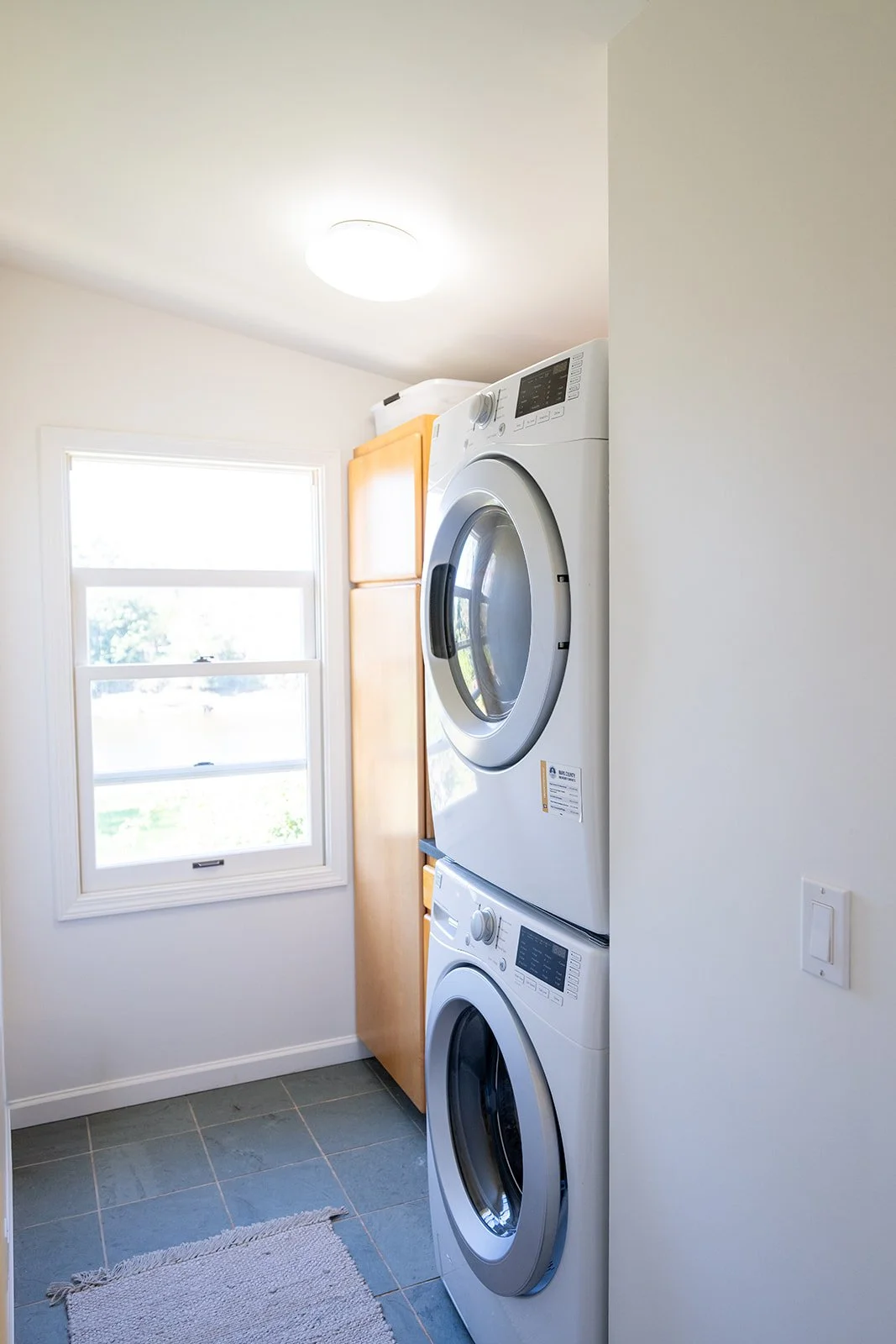 A laundry room with a stackable washer and dryer, a window, and a ceiling light.