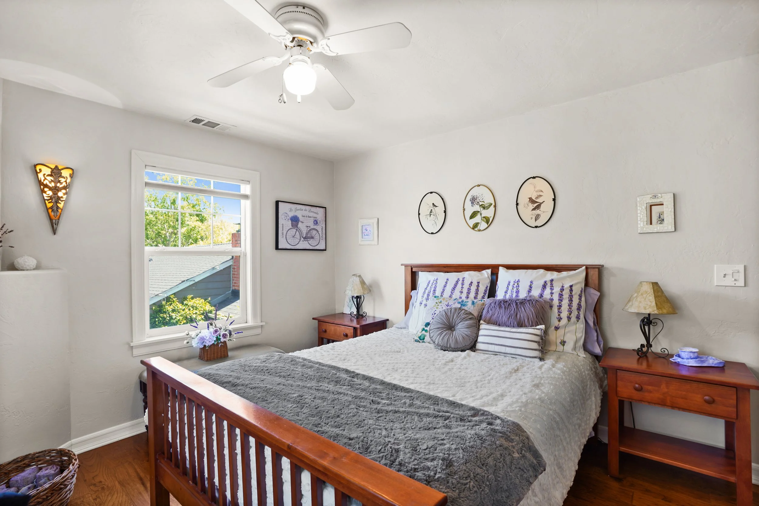 A cozy bedroom with a wooden bed, white bedding with lavender accents, two matching wooden nightstands, lamps, framed artworks and a ceiling fan, illuminated by natural light from a window showing trees outside.