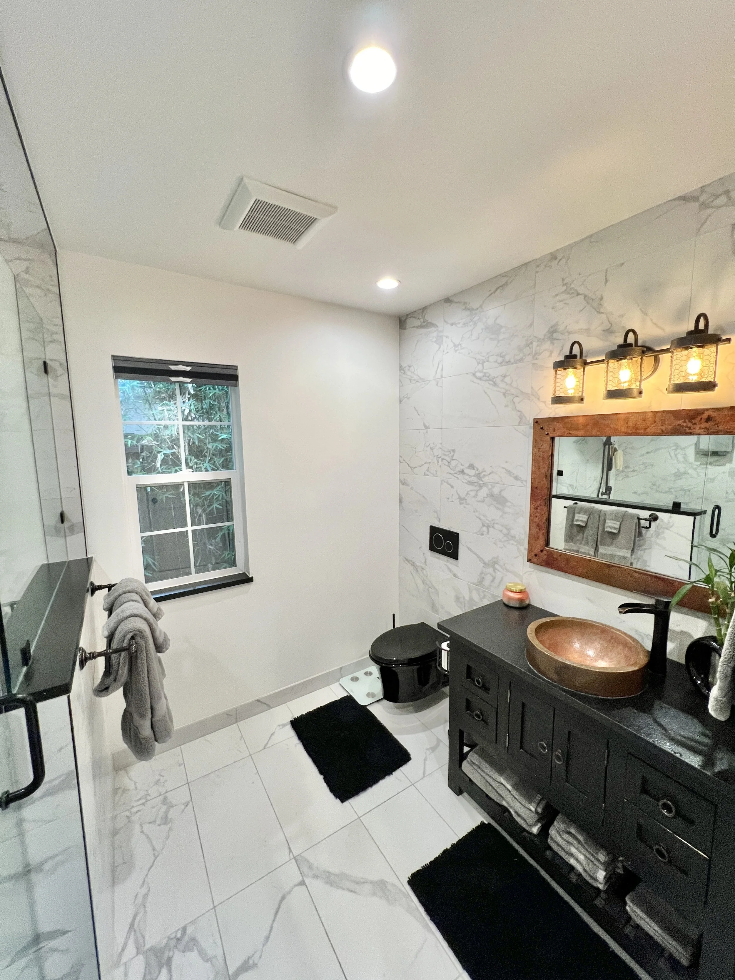 Modern bathroom with white marble tiles, black vanity with copper sink, large mirror with wooden frame, black toilet, black rugs, gray towels, window with a view of greenery, and decorative lighting above the mirror.
