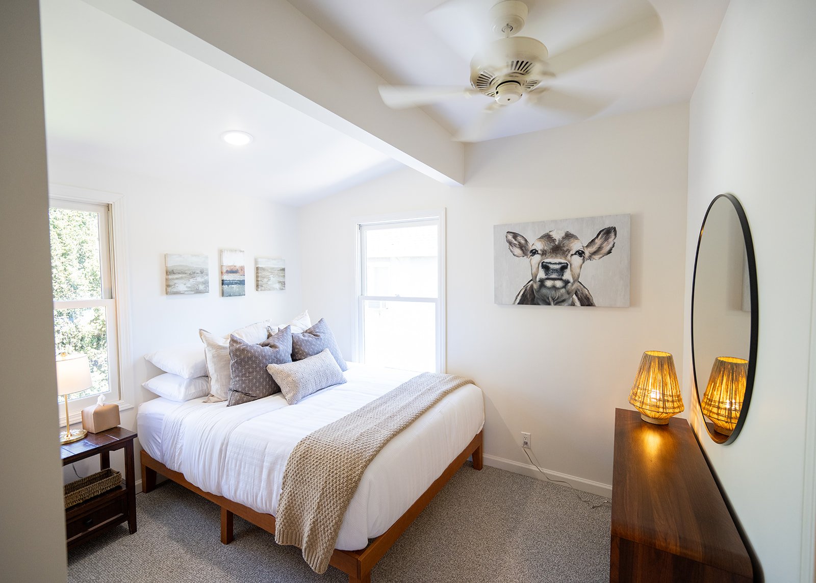 A bedroom with a white bed, several pillows, and a beige blanket. There is a nightstand with a lamp and a small object, and two windows providing natural light. The walls have three small landscape paintings and a large painting of a cow on the wall.
