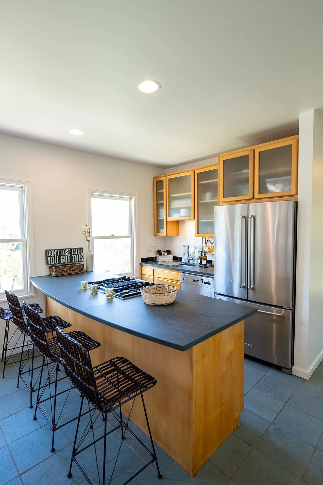 Modern kitchen with wooden cabinets, stainless steel refrigerator, black countertop, island with barstools, and windows letting in natural light.