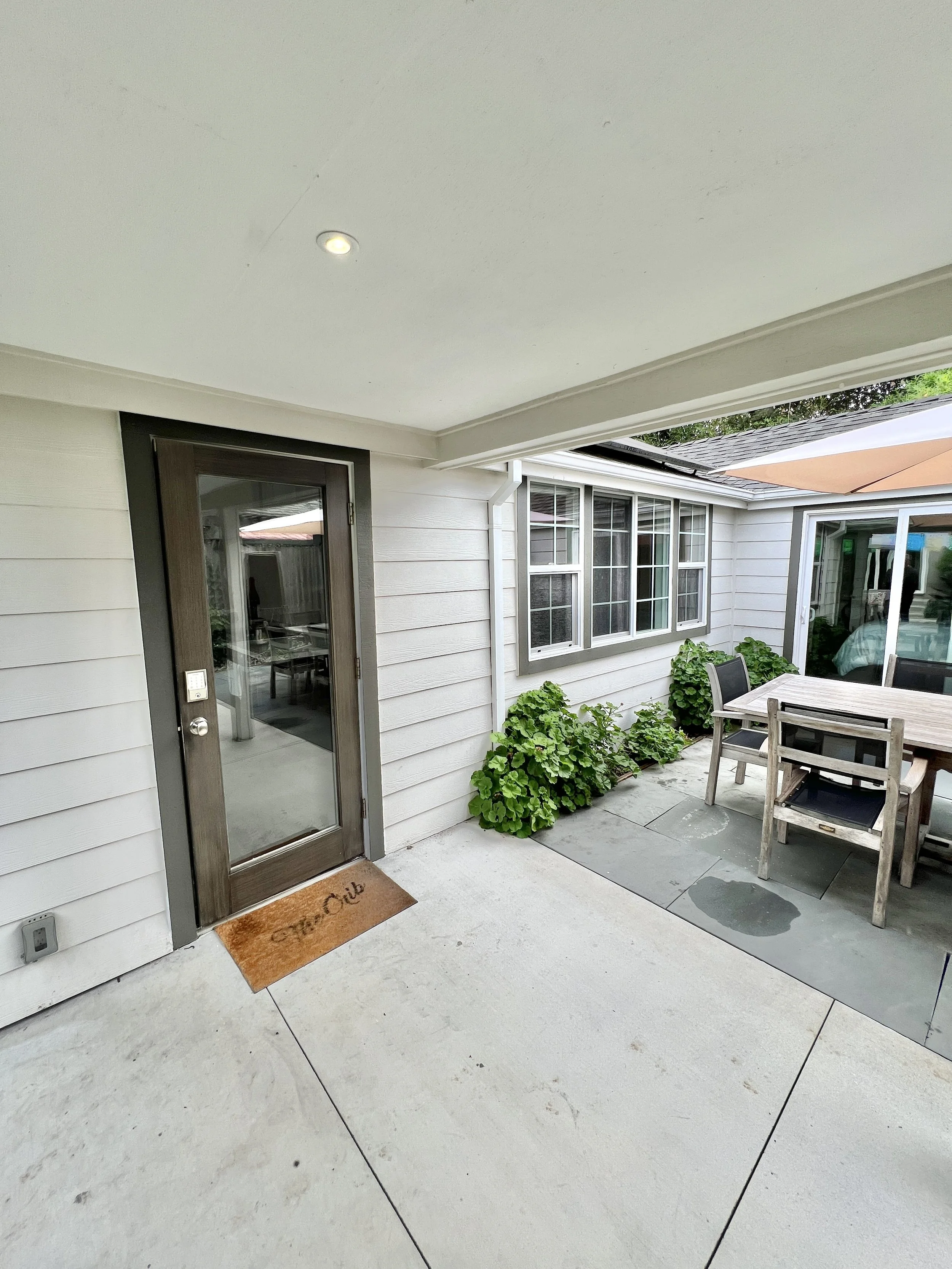 Covered backyard patio with wooden dining table and chairs, umbrellas, green plants, and house windows.
