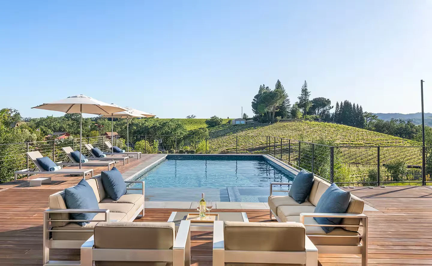 Luxury outdoor pool area with lounge chairs, umbrellas, and seating, overlooking green vineyards and hills under a clear sky.