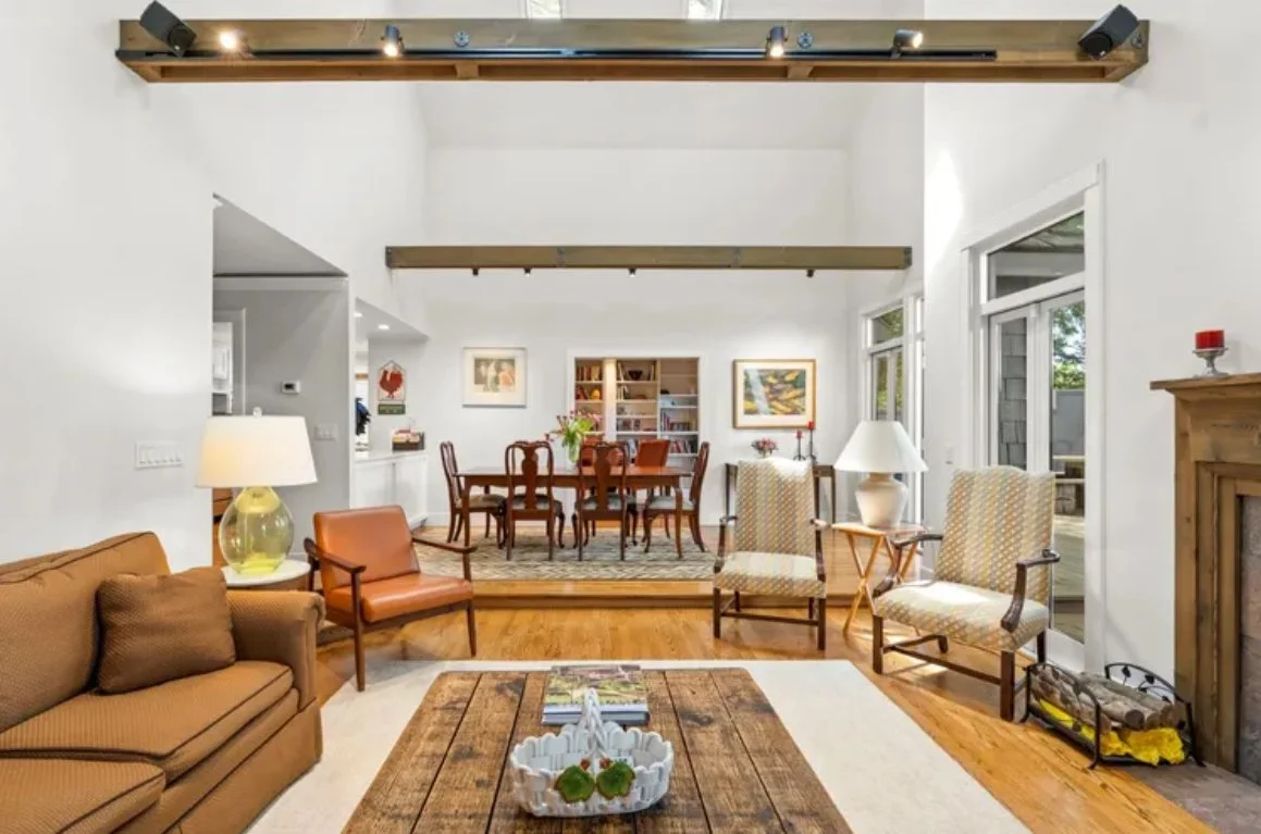 Living room with beige sofa, two armchairs, a coffee table, and a wooden fireplace. Dining area with a wooden table and six chairs, bookshelves, and artwork on the walls. Large windows and sliding door letting in natural light. Ceiling beams and a wo