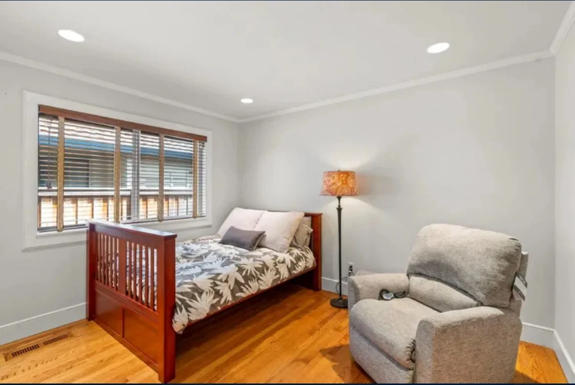 A small bedroom with a single bed, a window with blinds, a floor lamp, and a gray armchair on a wooden floor.