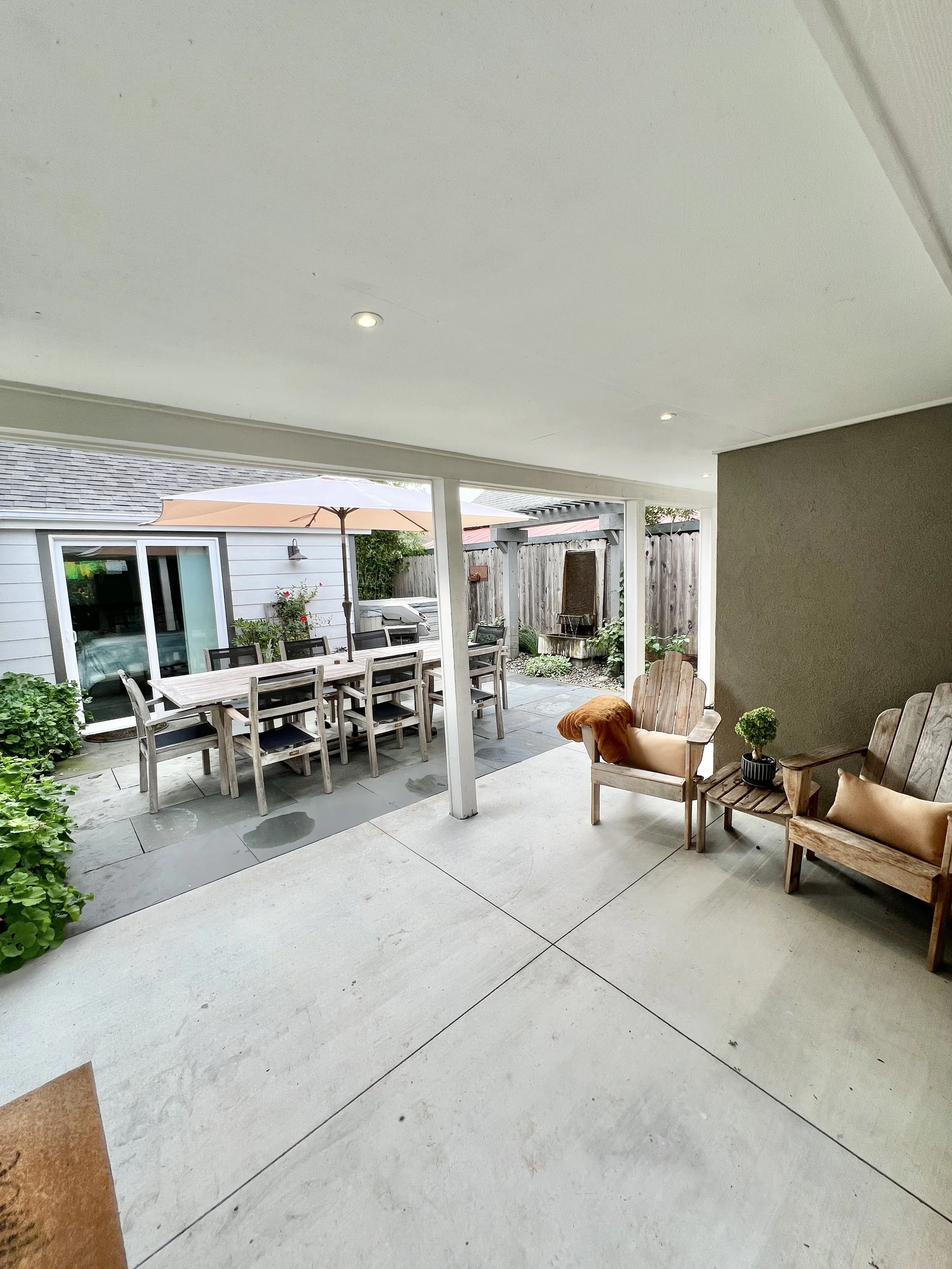 View of a covered patio area opening to an outdoor dining space with a table and chairs, and a separate seating area with wooden chairs, cushions, and plants.