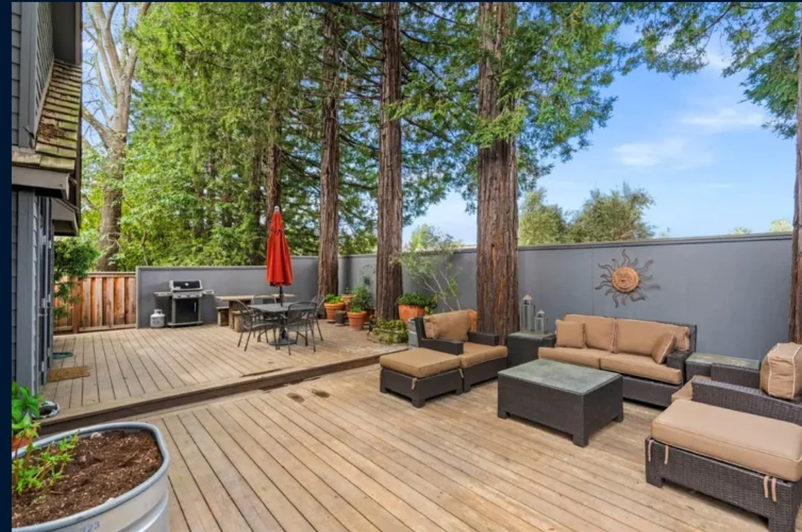 An outdoor wooden deck with patio furniture, including a sofa, chairs, and a coffee table, surrounded by tall trees and a gray fence with a sun wall decoration.