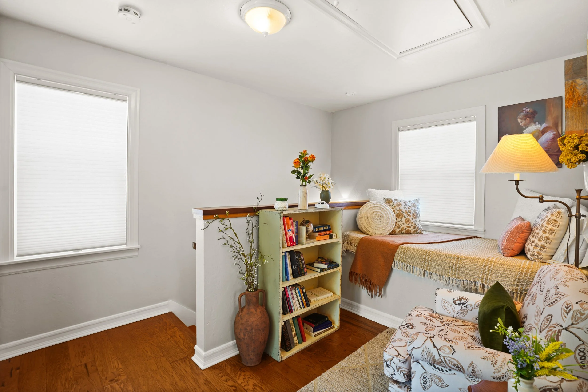 A cozy living room with white walls, two windows with blinds, a small bookshelf filled with books, a vase with orange flowers, a table lamp, and a sectional sofa with patterned and solid cushions. There is a painting of a woman on the wall.