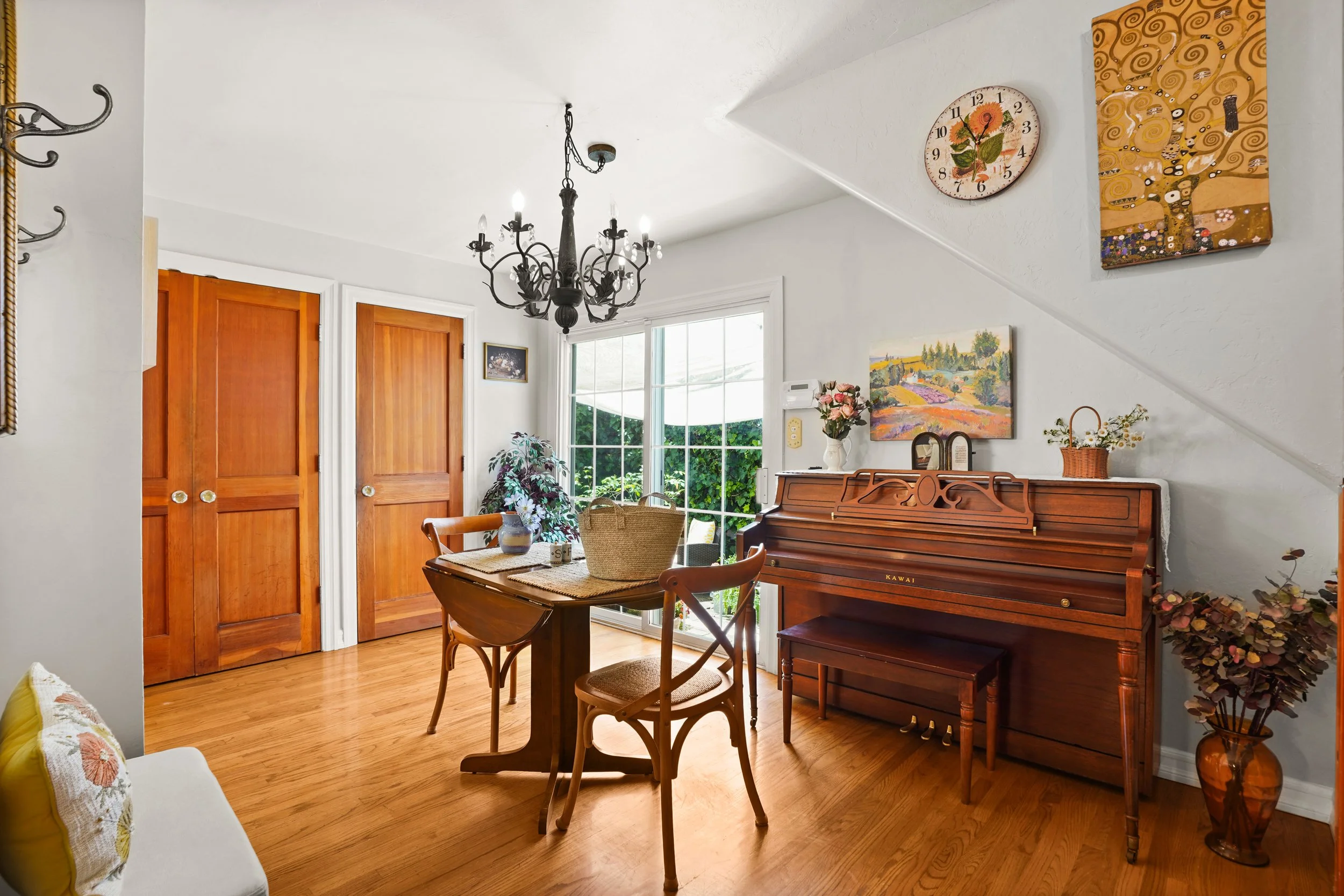 Interior of a living room with a wooden dining table, chairs, a vintage upright piano, and wall art. Sunlight streams through a large window, and there are plants and decorative items around.