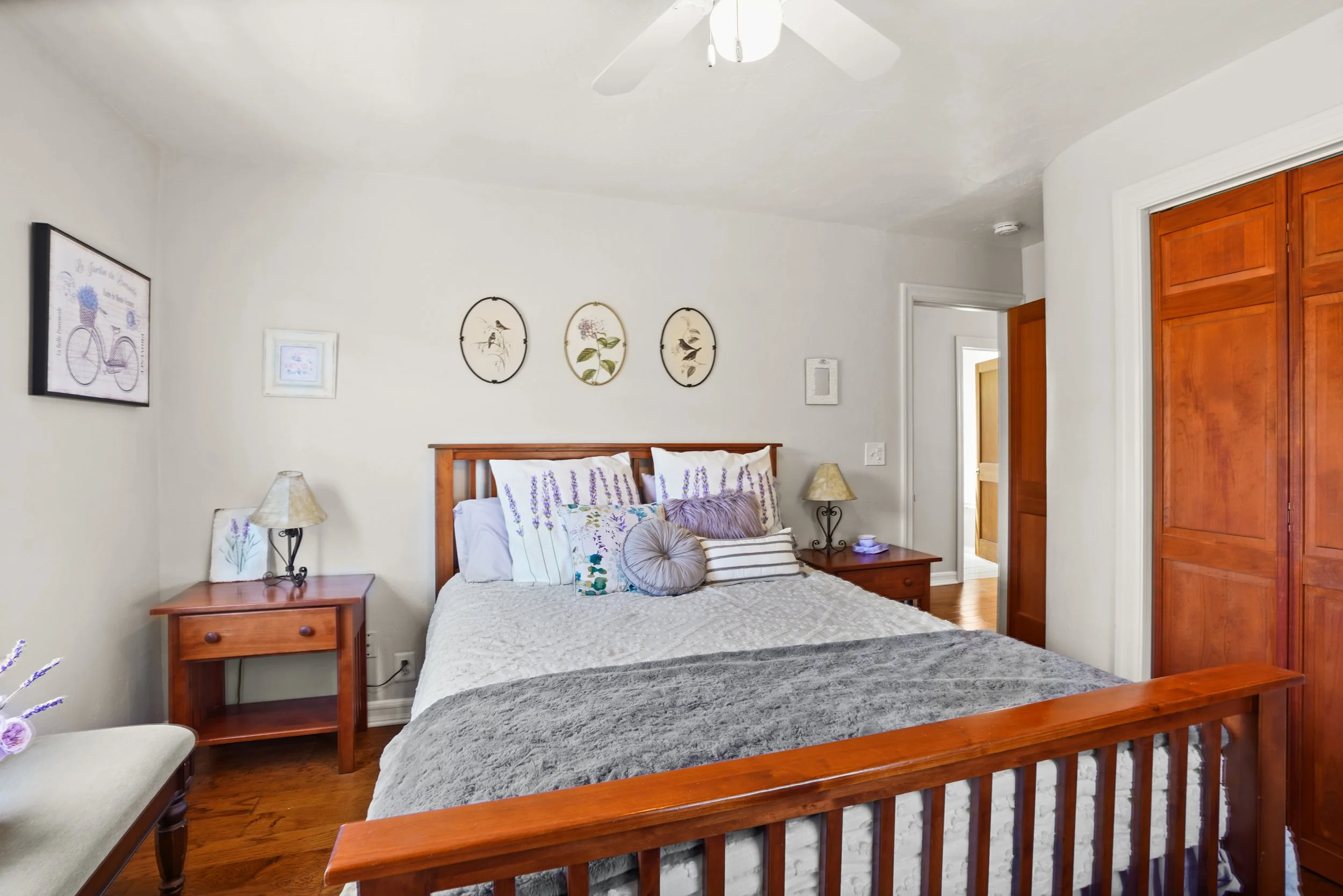 A bedroom with a wooden bed frame, white bedding, and decorative pillows. There are two wooden nightstands with lamps on either side of the bed. The wall above the bed has framed wall art, including three oval prints with birds and flowers. A ceiling