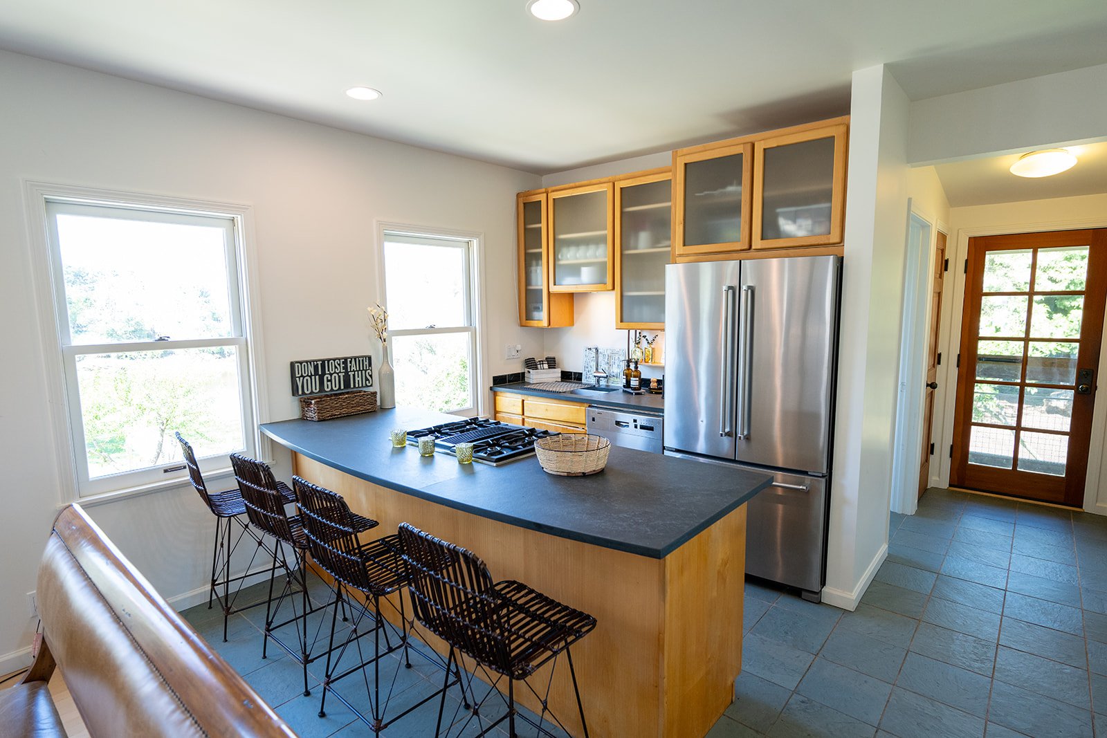 Kitchen with wooden cabinets, stainless steel refrigerator, black countertop, and barstools at a breakfast bar. Large windows with outside view, and a wooden door leading outside.