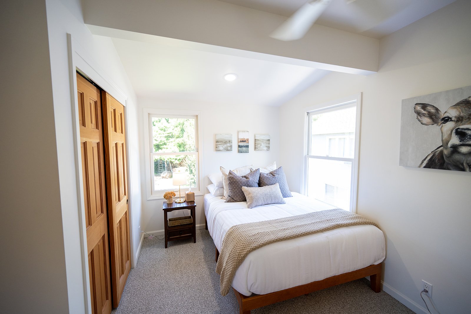 A bedroom with white walls, a bed with white bedding and gray pillows, a small wooden nightstand with a lamp and decor, two windows letting in natural light, a closet with wooden double doors, and artwork on the walls including a painting of a dog's 