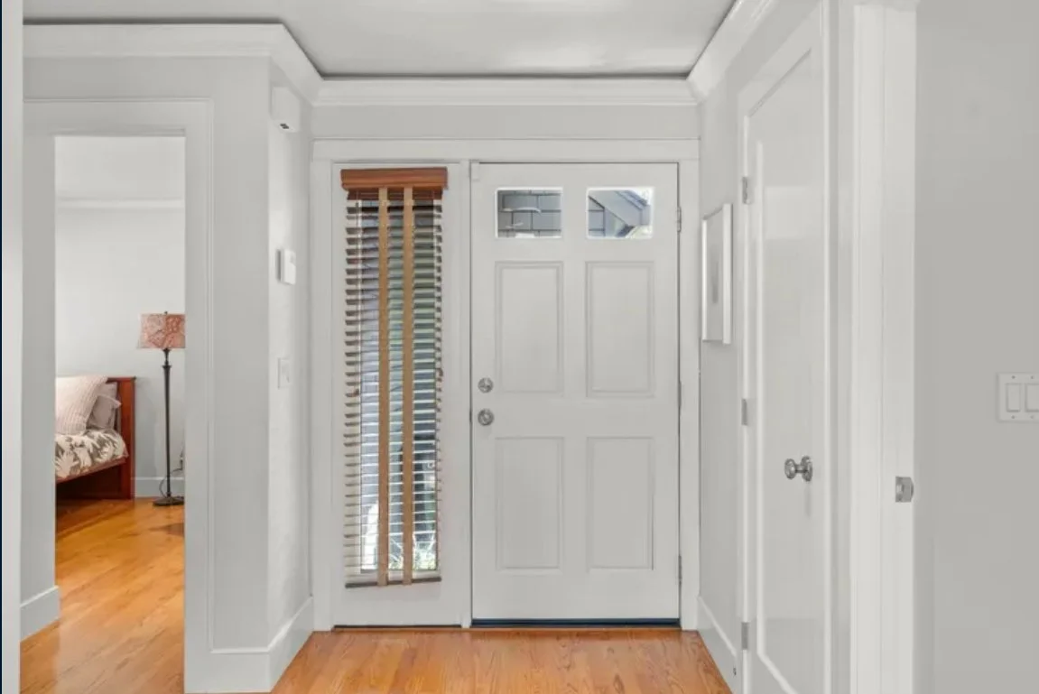 Interior view of a house entrance with a white front door, a glass panel, and a wooden door with blinds, leading to a bedroom with a bed and a nightstand with a lamp.
