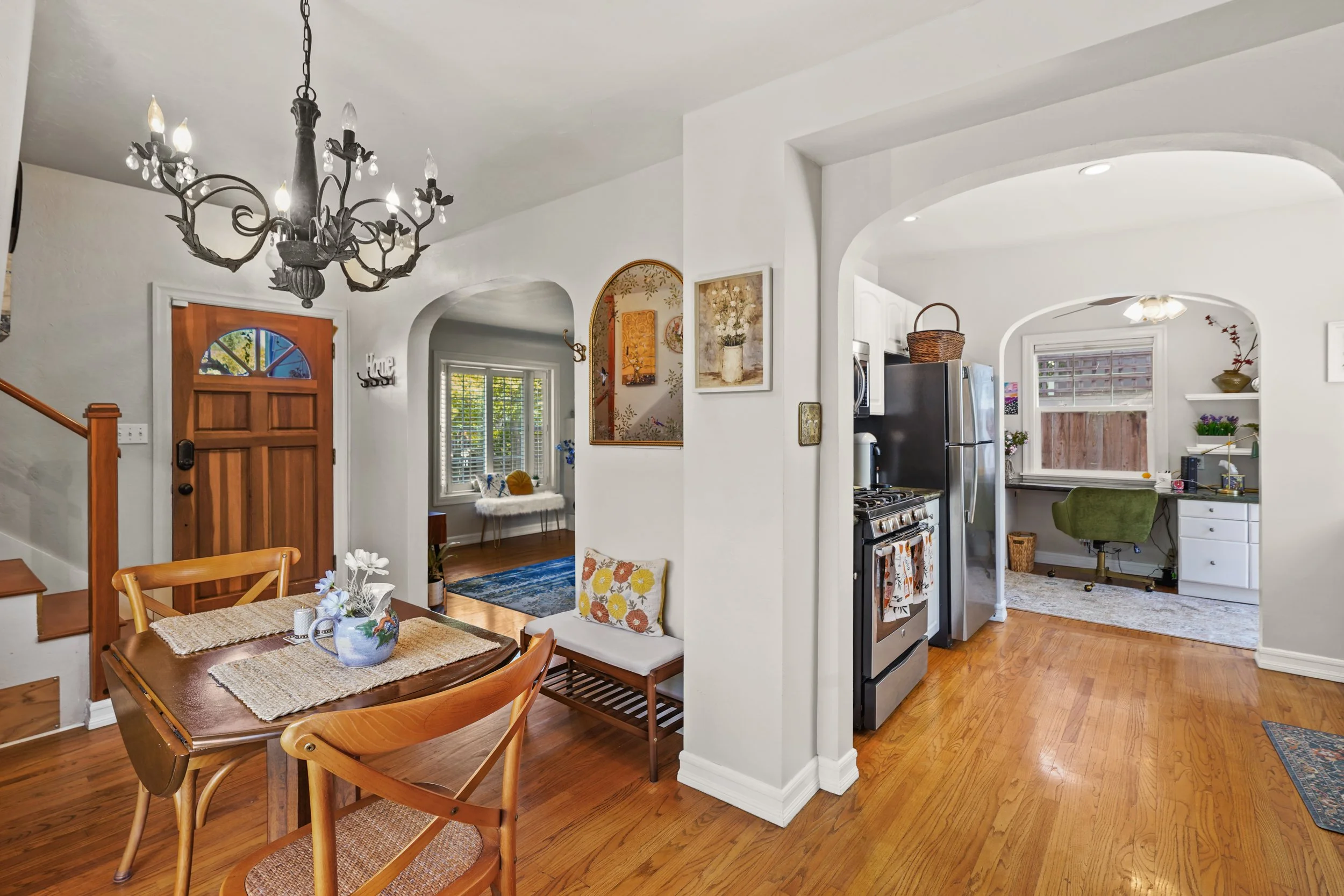 Interior of a home showing a dining area and kitchen with wooden floors, white walls, and various decorations, including a chandelier, paintings, and a decorative pillow on a corner bench.