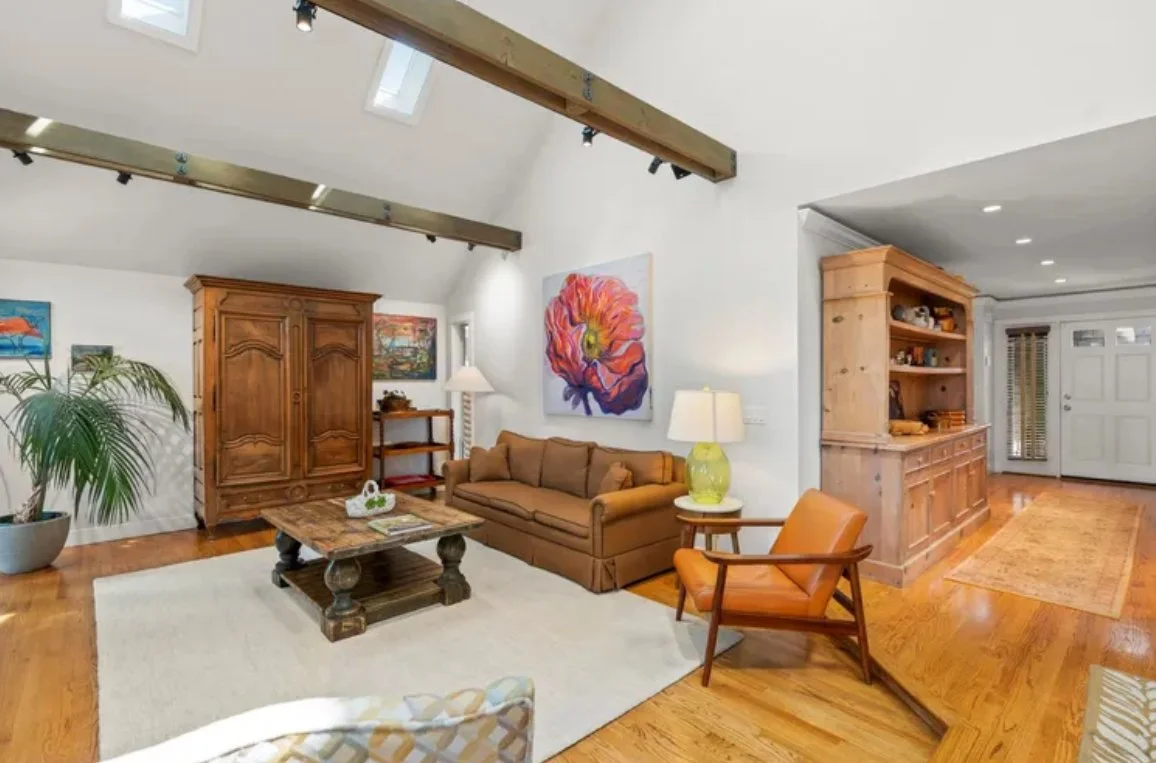 Living room with a brown sofa, orange armchair, wooden coffee table, large wooden armoire, wooden bookshelf, paintings on the wall, potted plant, and ceiling beams.