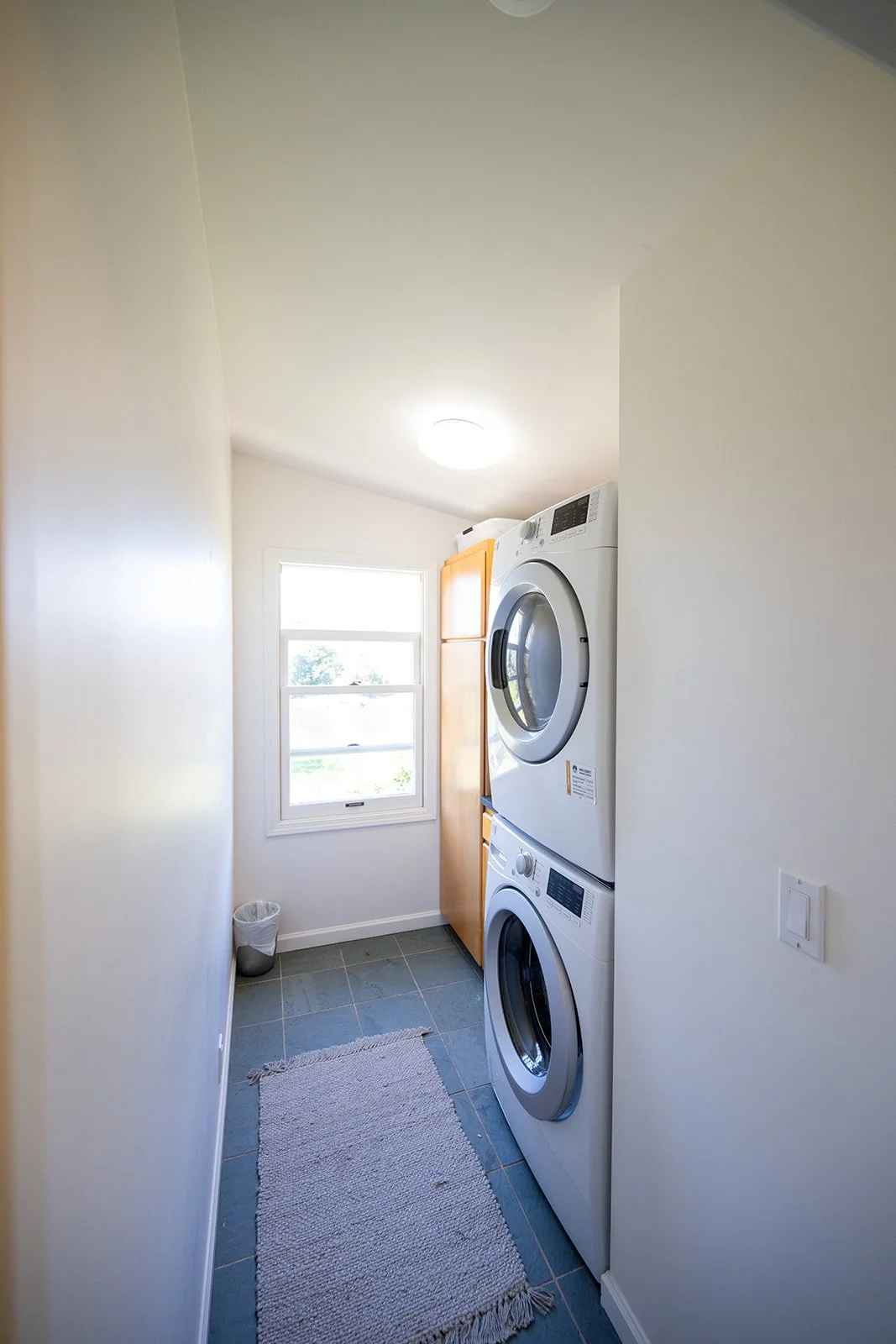Small laundry room with white walls, window, and stacked washer and dryer. Wooden cabinet, trash can, gray rug, and light switch visible.