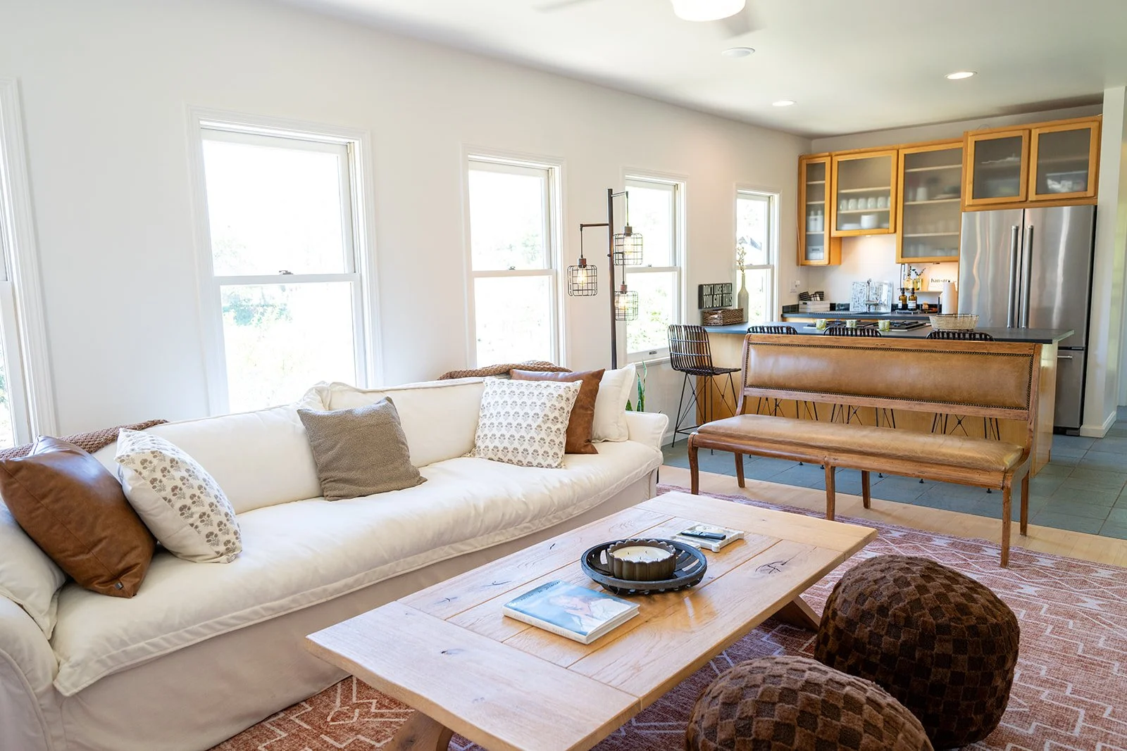 Bright living room with white couch and multiple pillows, wooden coffee table with books and a candle, two brown patterned poufs, open kitchen with light wood cabinets and stainless steel appliances, bar stools, hanging pendant lights, and large wind
