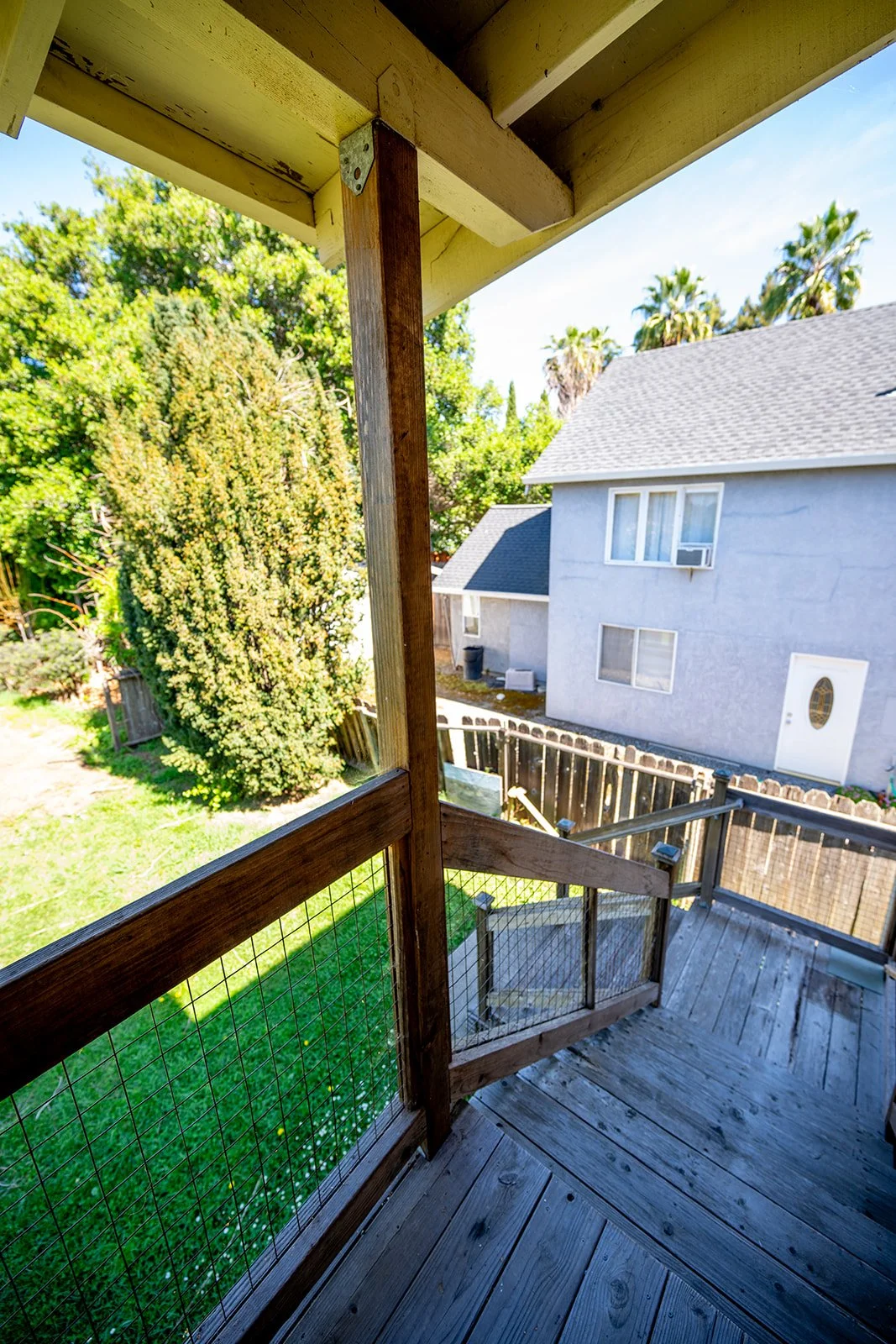 View from a wooden balcony with a wire railing, overlooking a backyard with a large bush and a neighboring house with gray exterior walls and windows, under a clear blue sky.