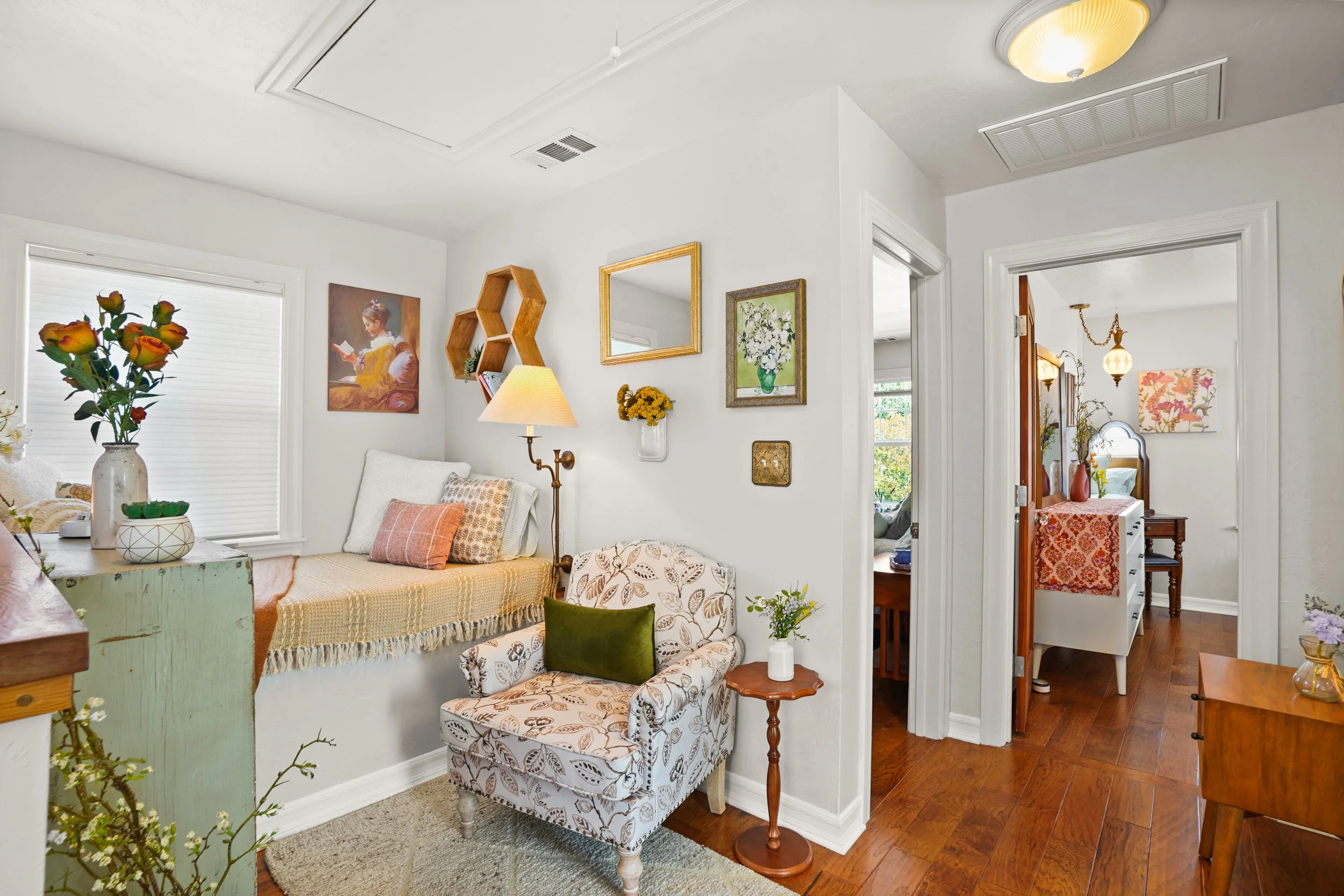 A cozy living room with white walls, wooden flooring, and decorated with framed and canvas art. There is a white loveseat with pillows, a floral armchair, a wooden side table with a white vase, and a standing lamp. The room has a window with closed b