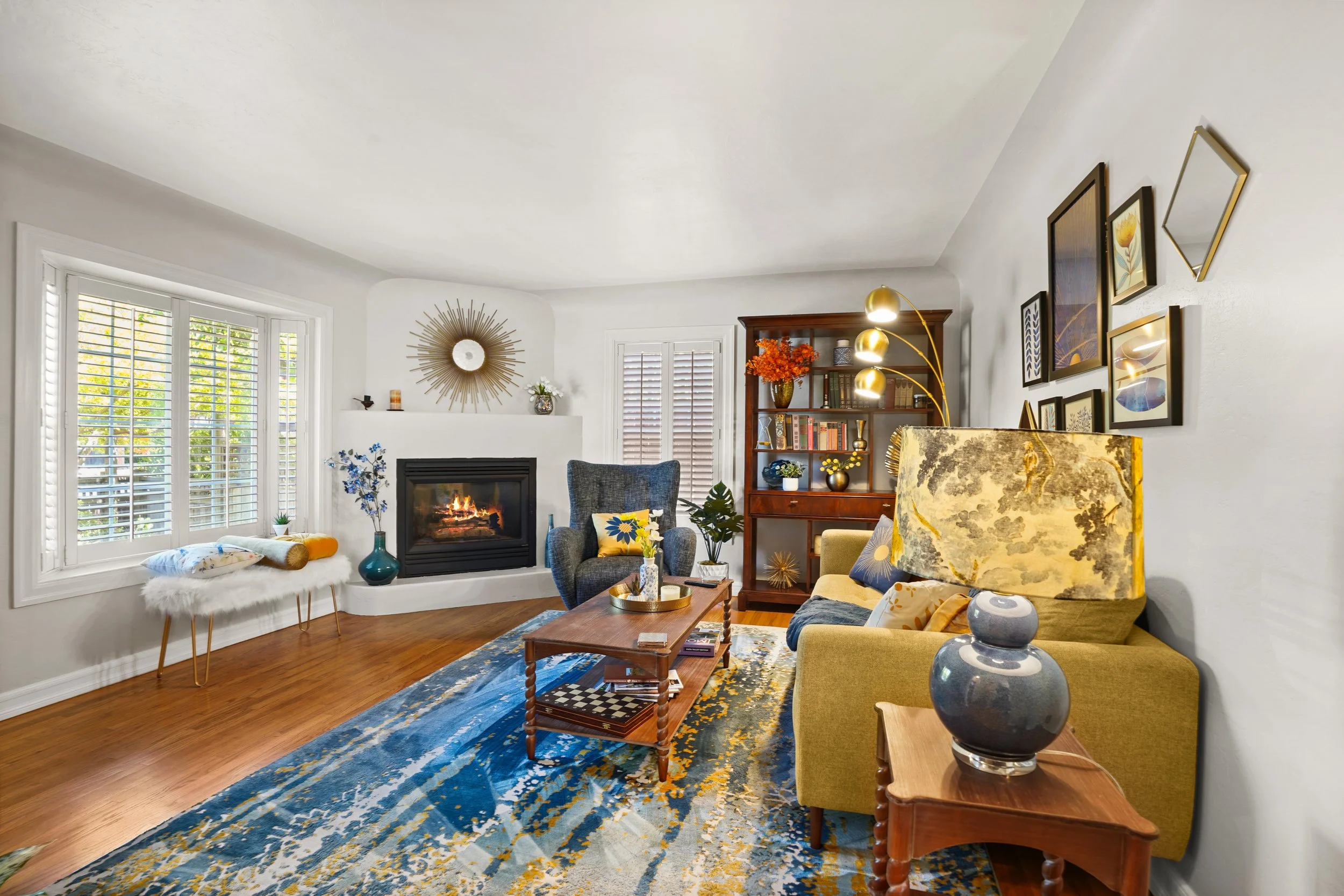 A living room with a white ceiling and walls, large bay window with shutters, a fireplace with decorative items on the mantel, a dark wooden bookshelf with books and decor, a cozy armchair, artwork and mirror on the walls, and a hardwood floor with a