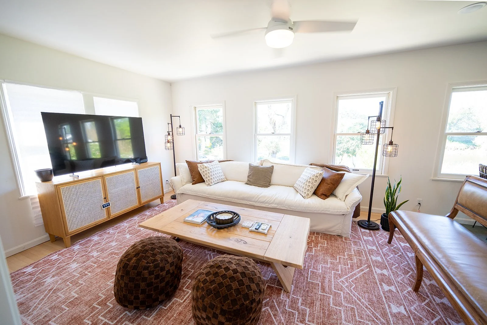Bright living room with white walls, large windows, a cream sofa with multiple pillows, a wooden coffee table, a TV on a wooden stand, a floor lamp, and a pink patterned rug.