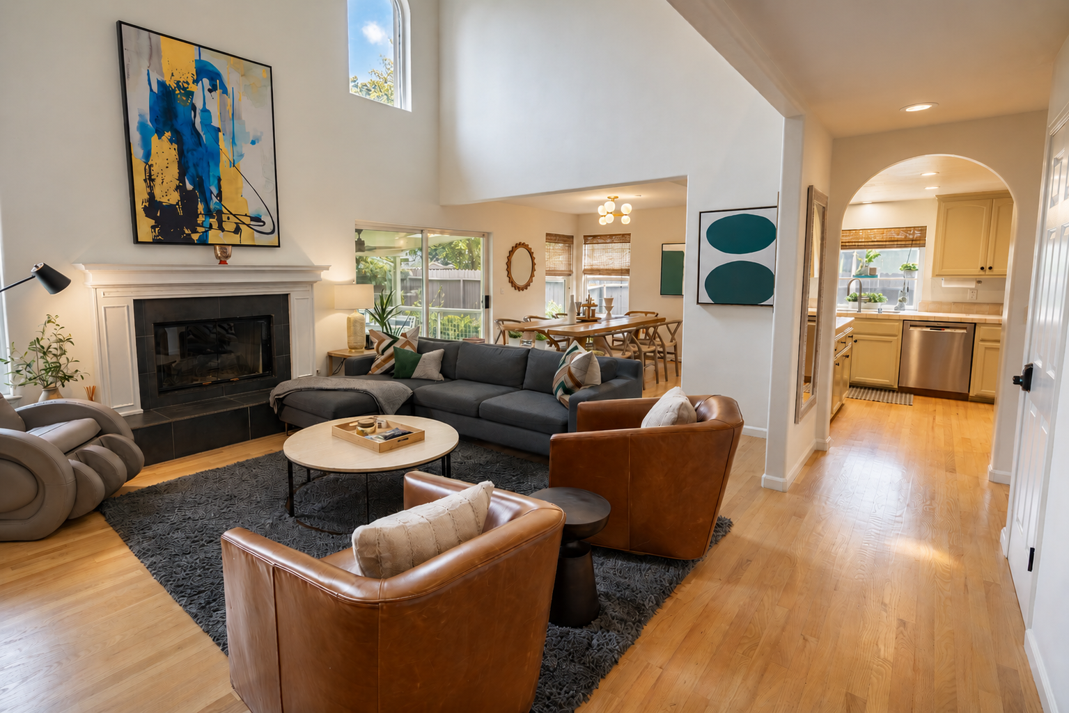 Living room with a gray sofa, leather armchairs, a fireplace, and artwork on the walls, connected to a dining area and kitchen with wooden floors and large windows.