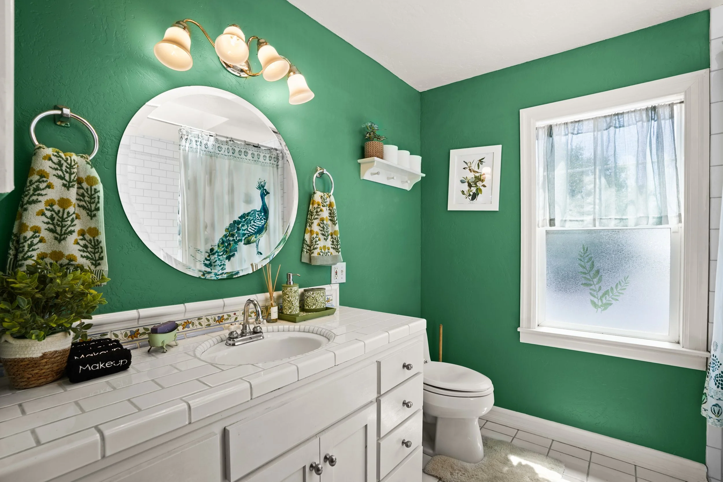 A bathroom with green painted walls, a white tile countertop with a sink, a round mirror, and a window with frosted glass and curtains. Decorative towels and plants are also present.
