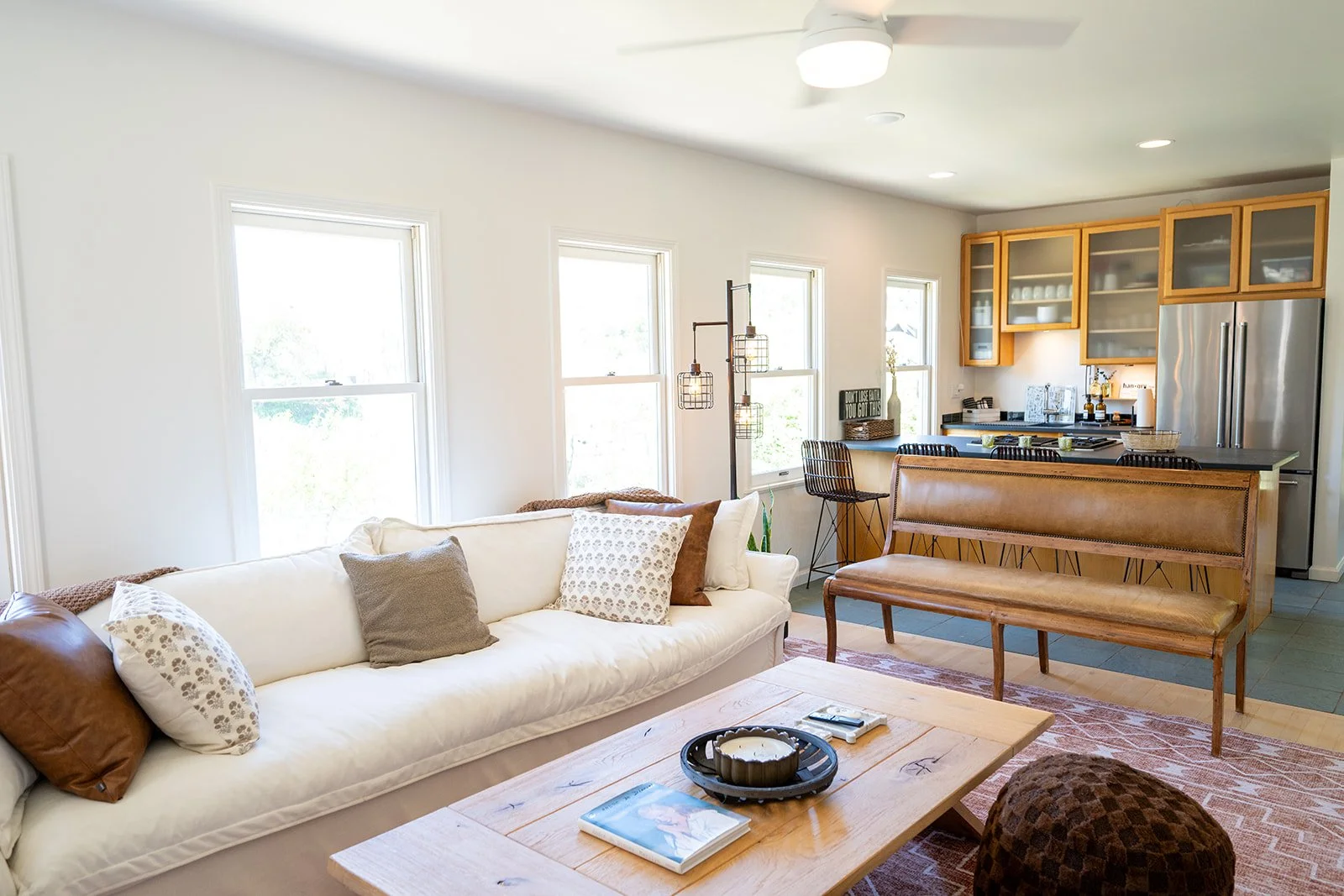 Living room with white couch, pillows, wooden coffee table, and kitchen with wooden cabinets, stainless steel refrigerator, and barstools.