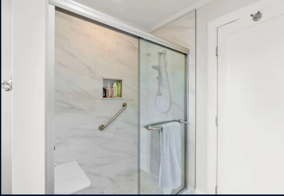 A glass shower with marble walls, a built-in shelf with toiletries, an adjustable showerhead, and a white towel hanging on the towel bar inside a bathroom.