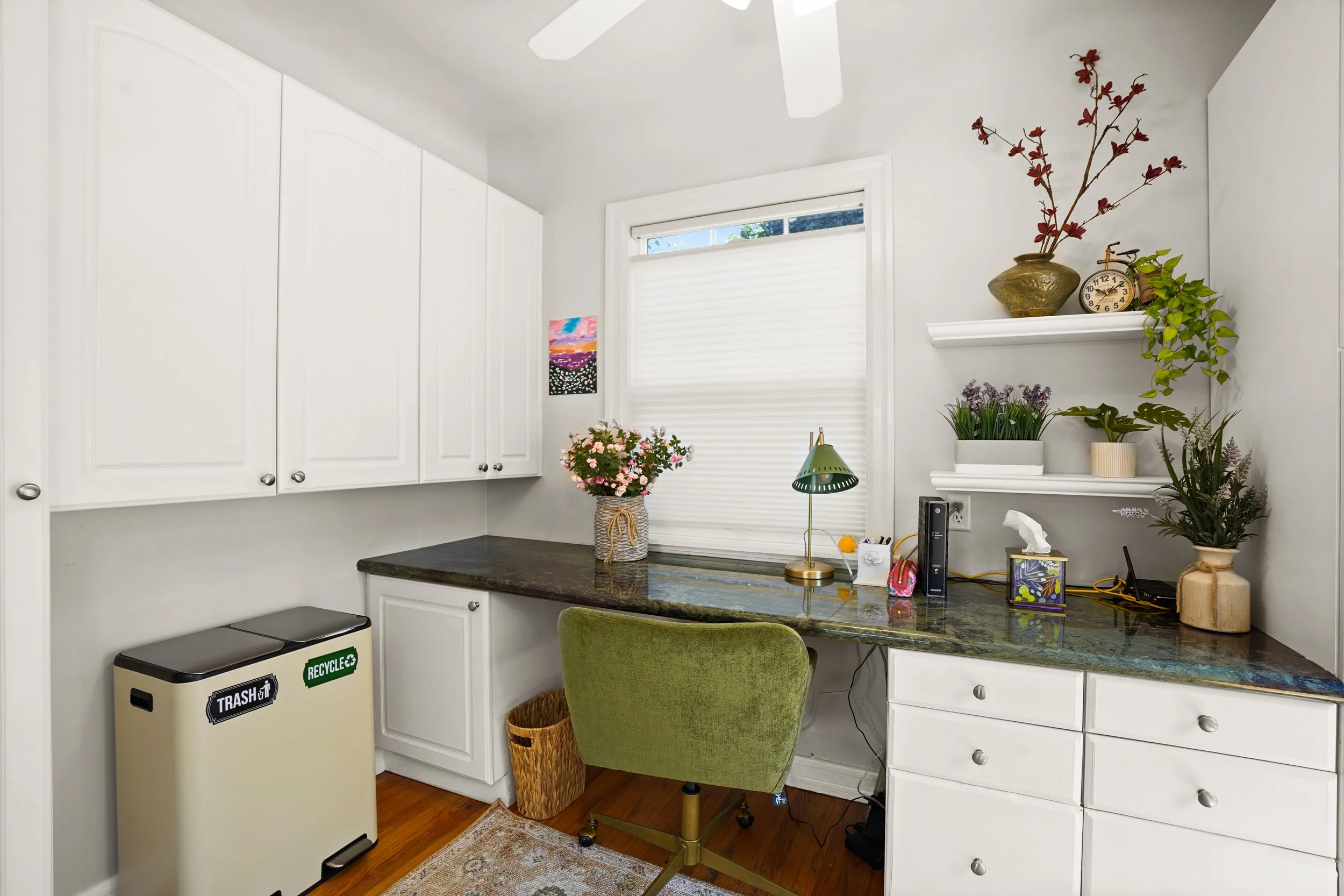 A home office corner with white cabinets, a window with blinds, a green chair, and various decorative plants and items on shelves.