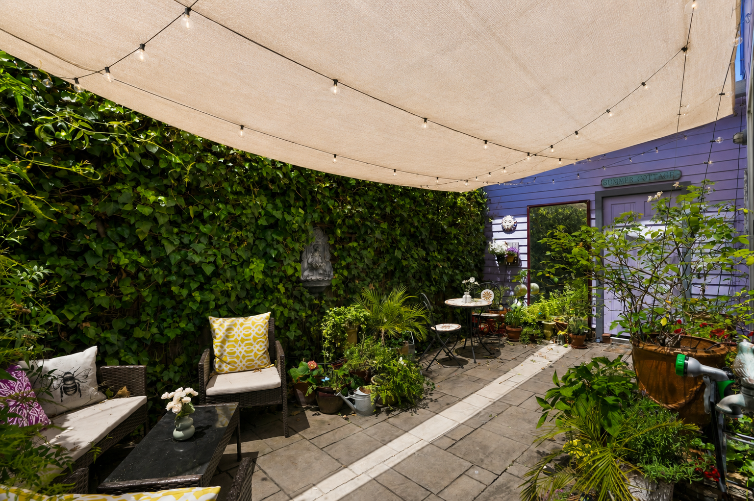 Vertical outdoor patio area with blue house wall labeled 'Summer Cottage,' surrounded by greenery, string lights, patio furniture with cushions, potted plants, and a shaded canopy.