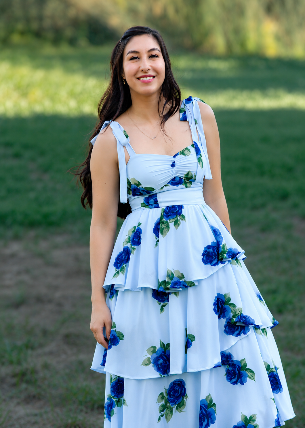 A young woman in a white dress with blue roses standing outdoors on grass, smiling at the camera with a background of trees.