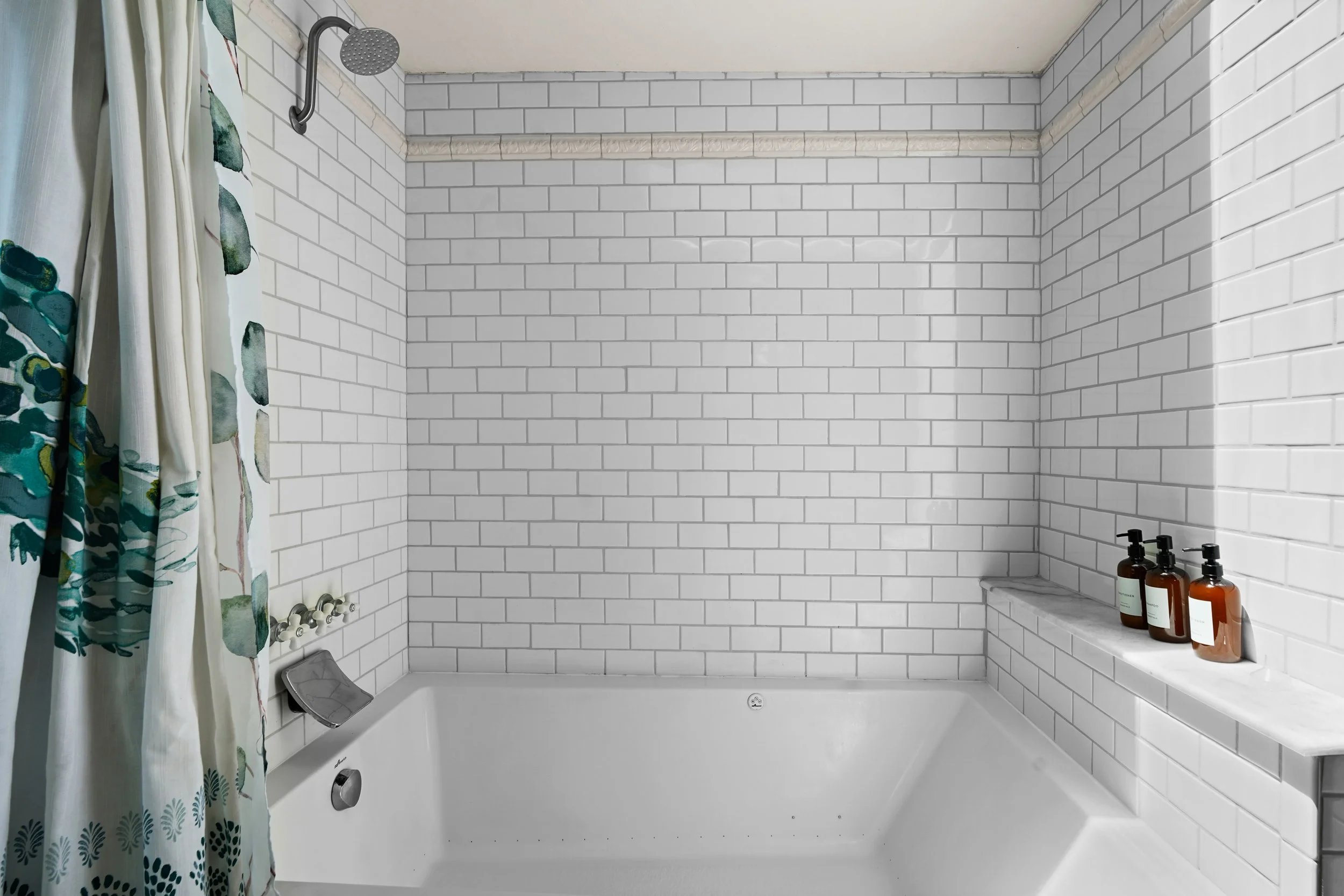 Bathroom with white subway tile walls, a bathtub with a shower, and a shelf with brown bottles. Shower curtain with a green and beige floral pattern hanging on the left. Showerhead mounted on the wall above the bathtub.
