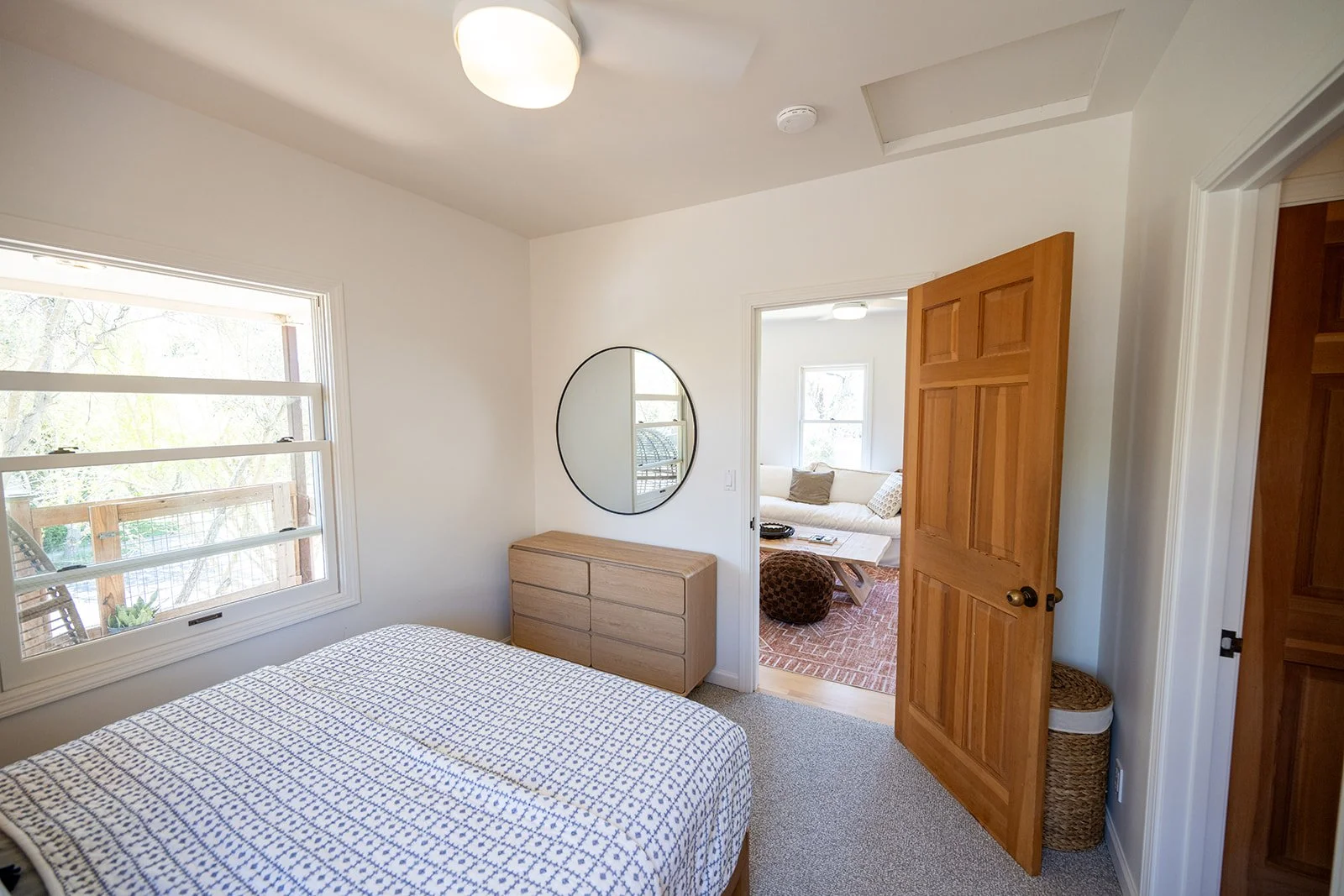 Bedroom with a bed, a window, a wooden dresser, a round mirror, and an open door leading to a living room with a couch and windows.