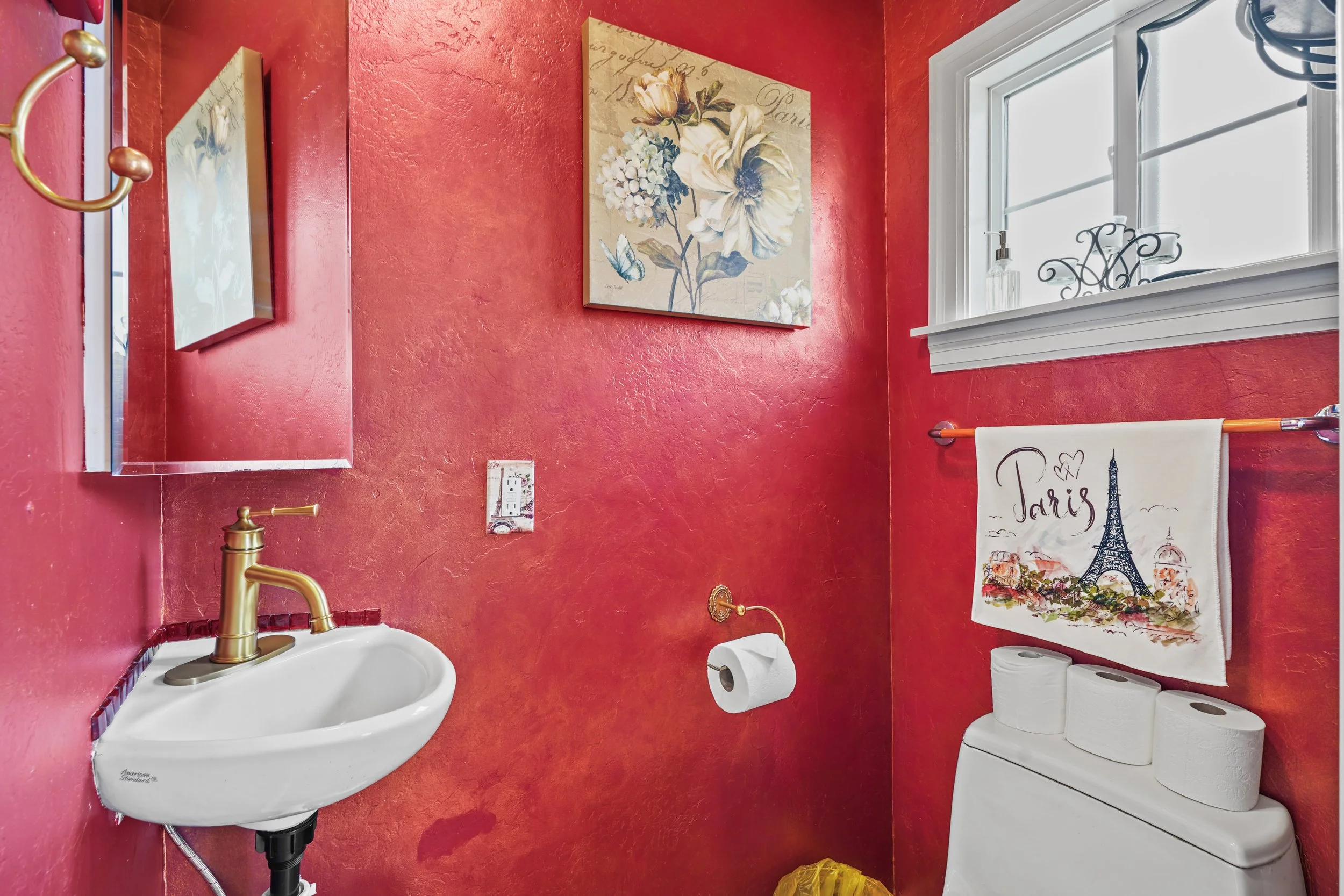 A small bathroom with red textured walls, a white sink with gold fixtures, a toilet, a mirror, a window with white trim, and artwork on the walls, including a floral painting and a Paris-themed towel with the Eiffel Tower.
