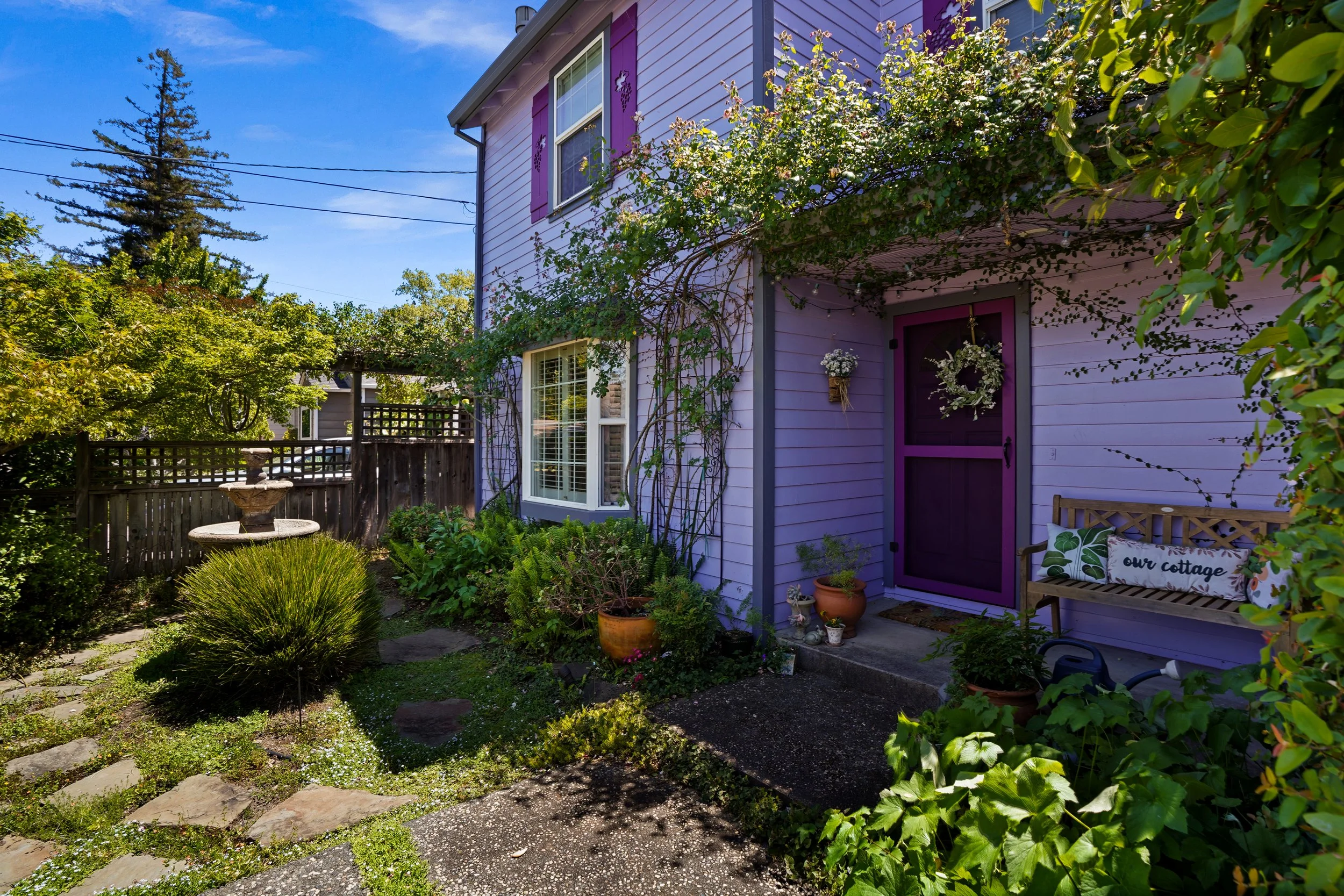 A purple house with a purple door, decorated with flower wreaths and surrounded by plants and trees, under a clear blue sky.