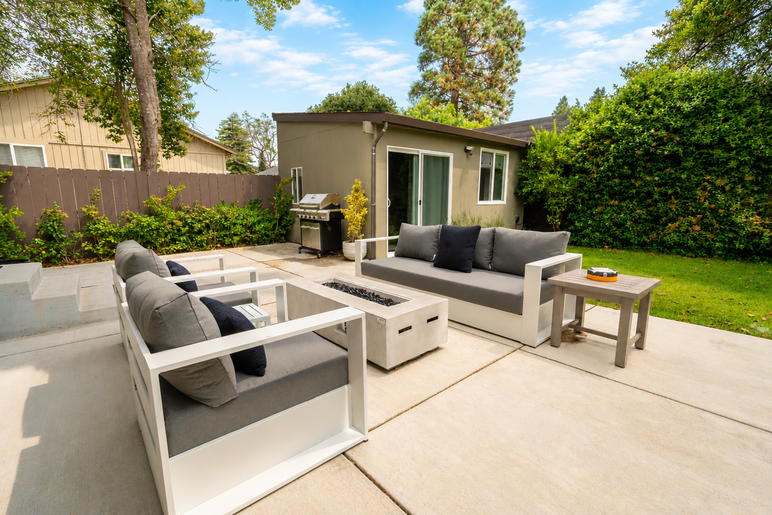 Outdoor patio with gray cushioned seating, a fire pit, barbecue grill, small side table, green bushes, tall trees, and a small green house with a sliding glass door and windows, under a clear blue sky.