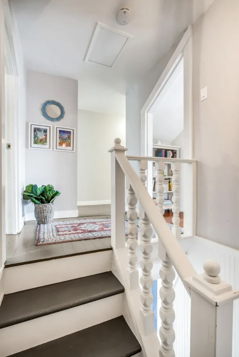 Interior view of a staircase landing with white wooden balusters, a patterned rug, a potted plant, and picture frames on the wall.