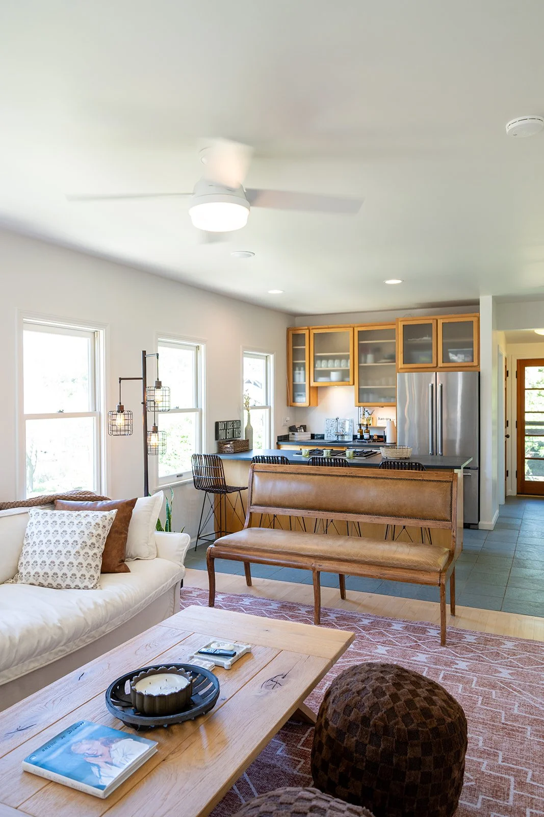 Living room and kitchen with white walls, three large windows, a white sofa with patterned pillows, a wooden coffee table with a magazine and candle, a brown upholstered chair, a patterned rug, a kitchen counter with black bar stools, wooden cabinets
