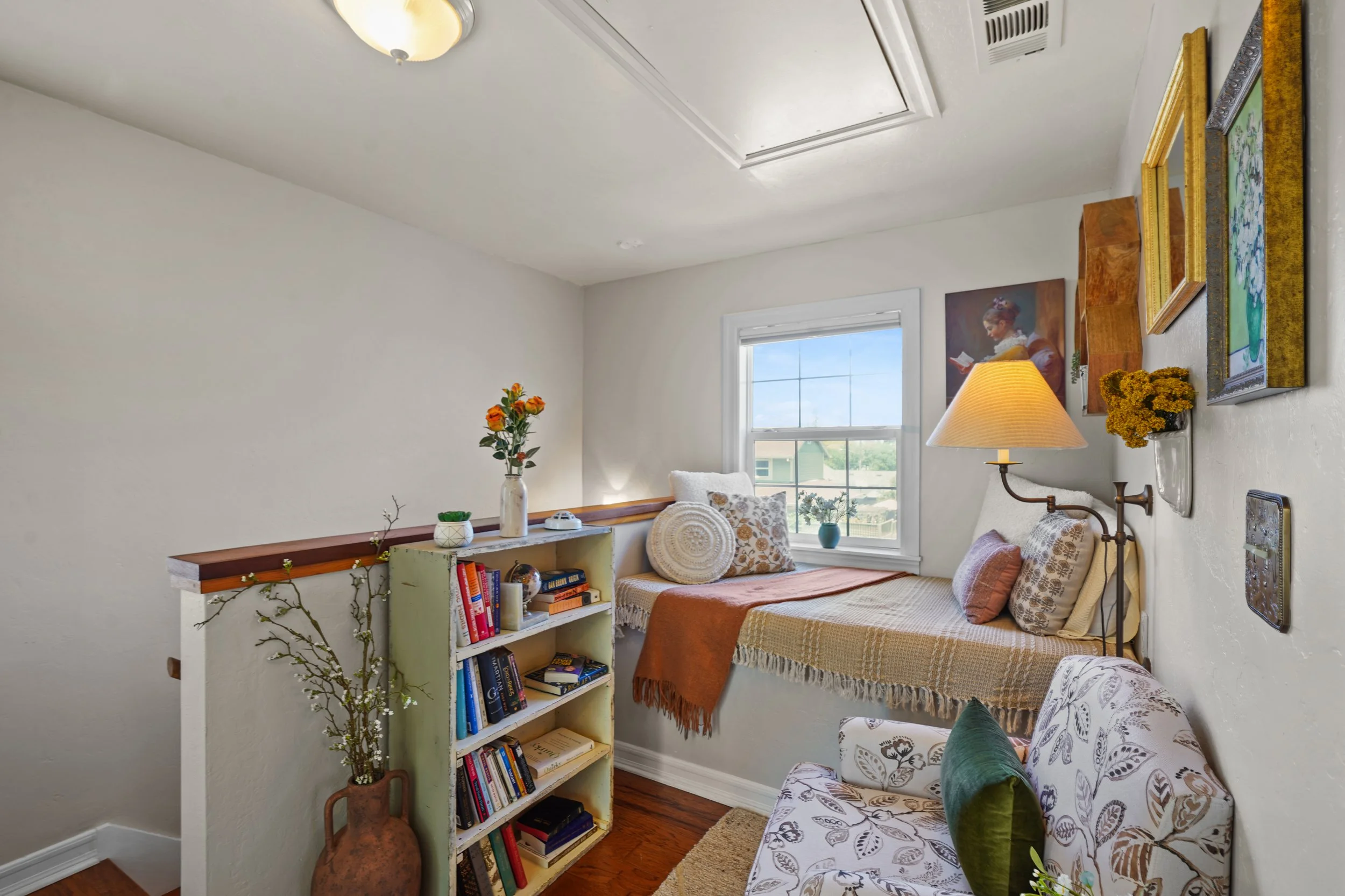 A cozy corner of a room with a window letting in natural light, decorated with paintings, a lamp, and plants. There is a bed with pillows and a bench with books and decor.