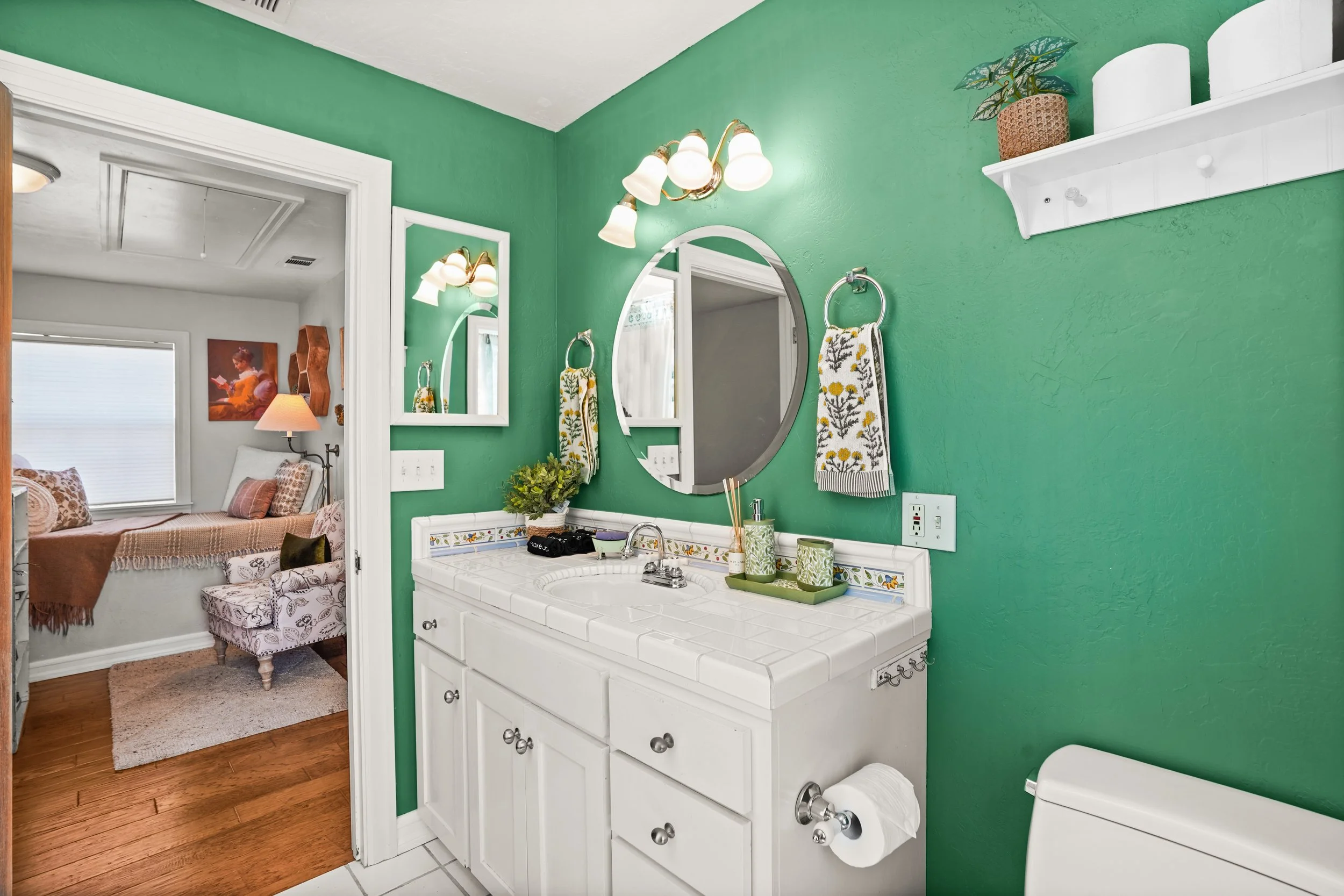 Bathroom with green walls, white vanity with tile counter, oval mirror, wall-mounted light fixture, plant on shelf, and towels hanging. Adjacent to a bedroom with sitting area, patterned chairs, artwork, window with blinds, and hardwood floors.