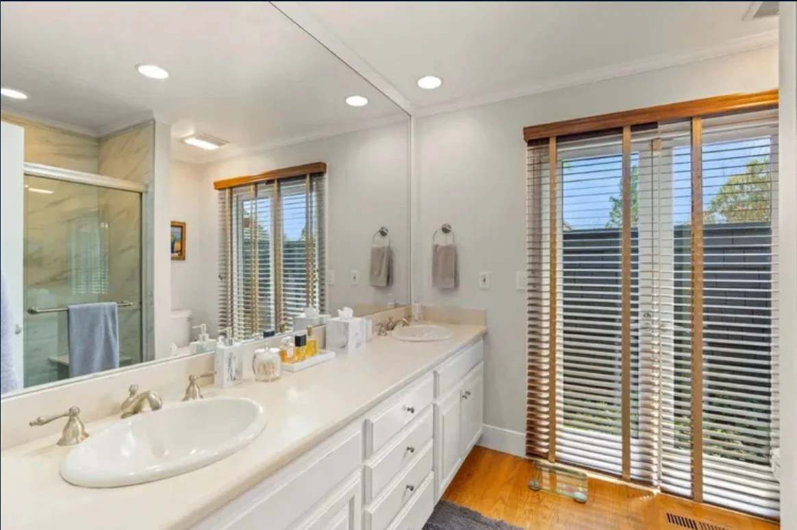 A spacious bathroom with a double sink vanity, large mirror, and wooden blinds on two windows. The room has white cabinets, a glass shower enclosure, and hardwood floors.
