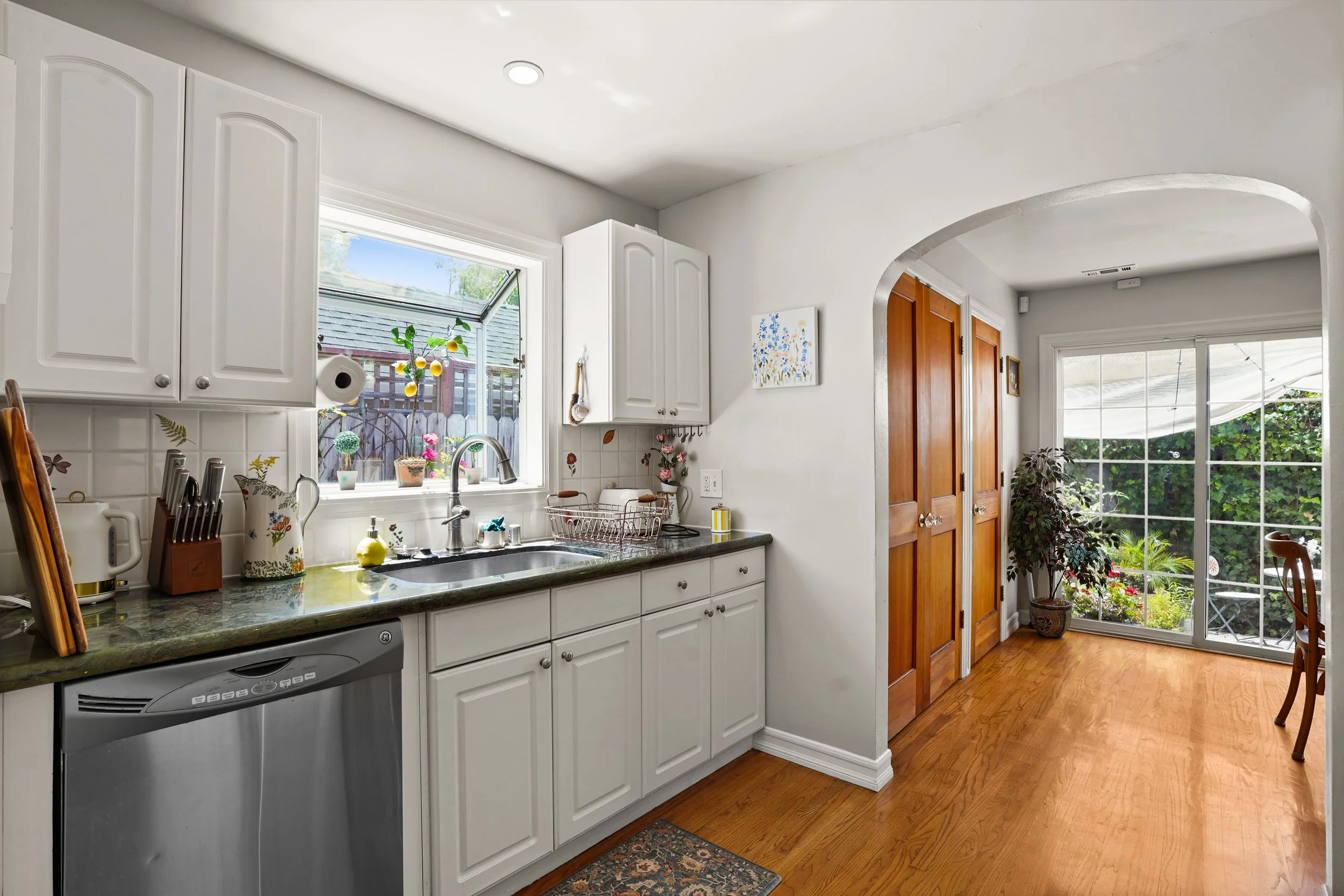 A clean kitchen with white cabinets, a granite countertop, a stainless steel dishwasher, a window above the sink showing a garden, and a view of hardwood flooring and a sliding glass door leading outside.