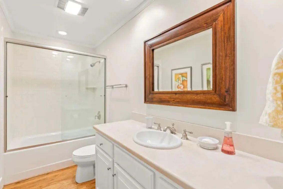Bathroom with a shower, toilet, sink, large wooden-framed mirror, and framed artwork on the wall.