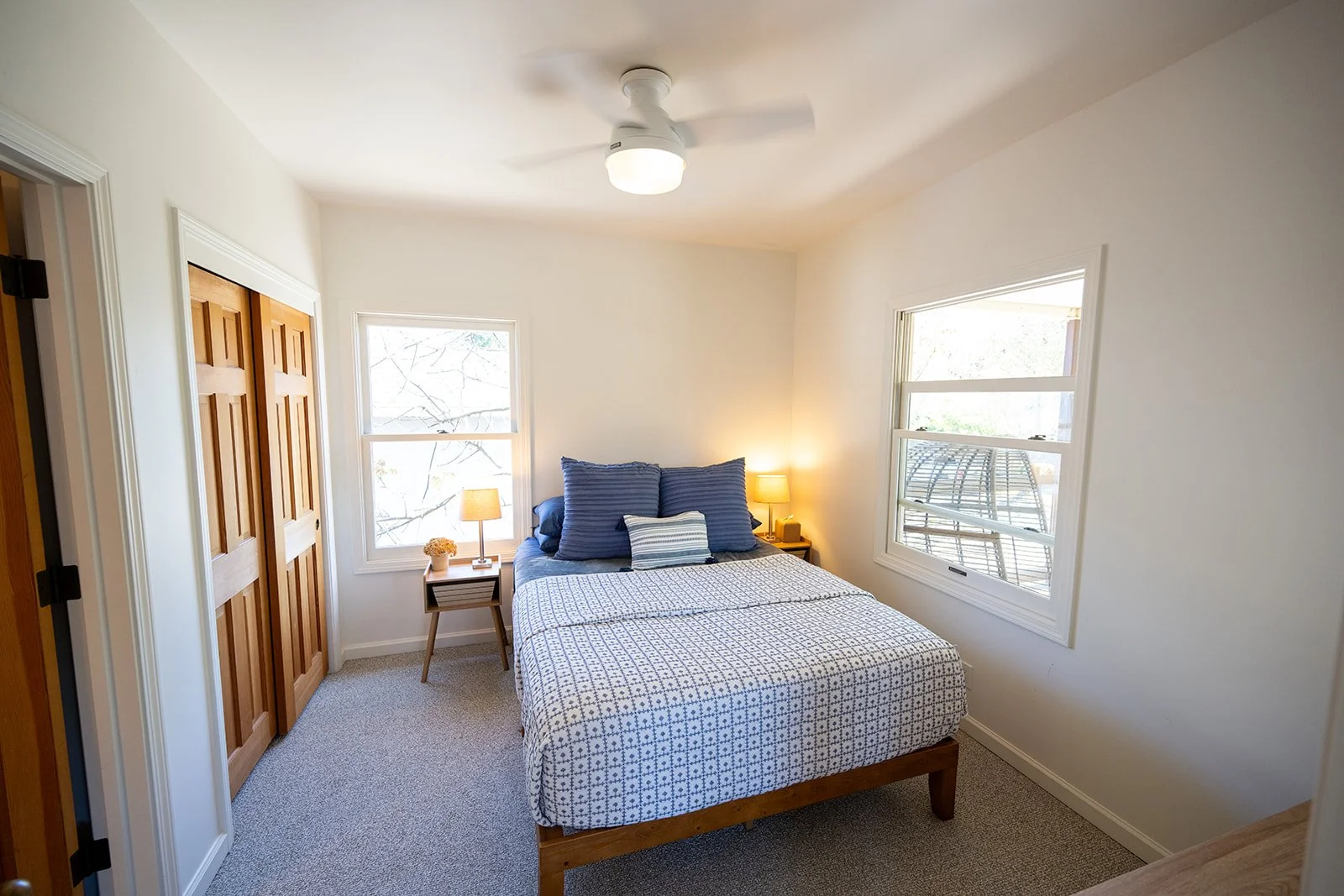 A bedroom with a bed, two windows, and a ceiling fan. The bed has a patterned bedsheet, multiple pillows, and two nightstands with lamps.