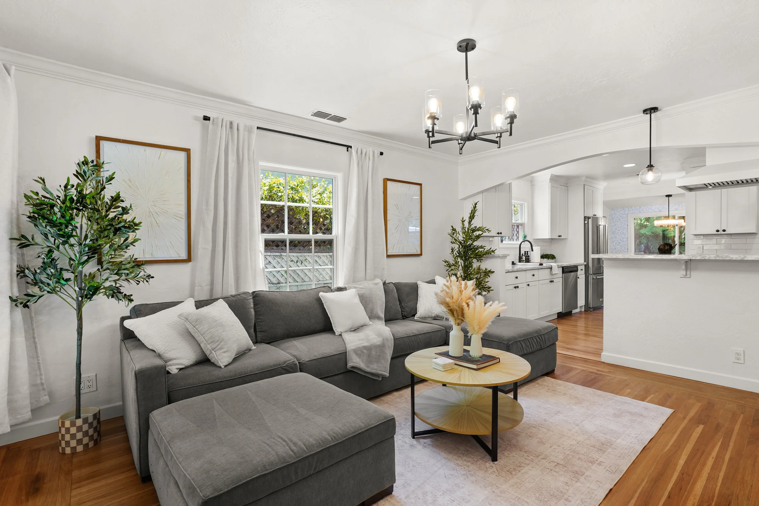 Living room with gray sectional sofa, white pillows, and a round coffee table with vases, adjacent to a kitchen with white cabinets and stainless steel appliances, featuring large windows with white curtains and hardwood floors.